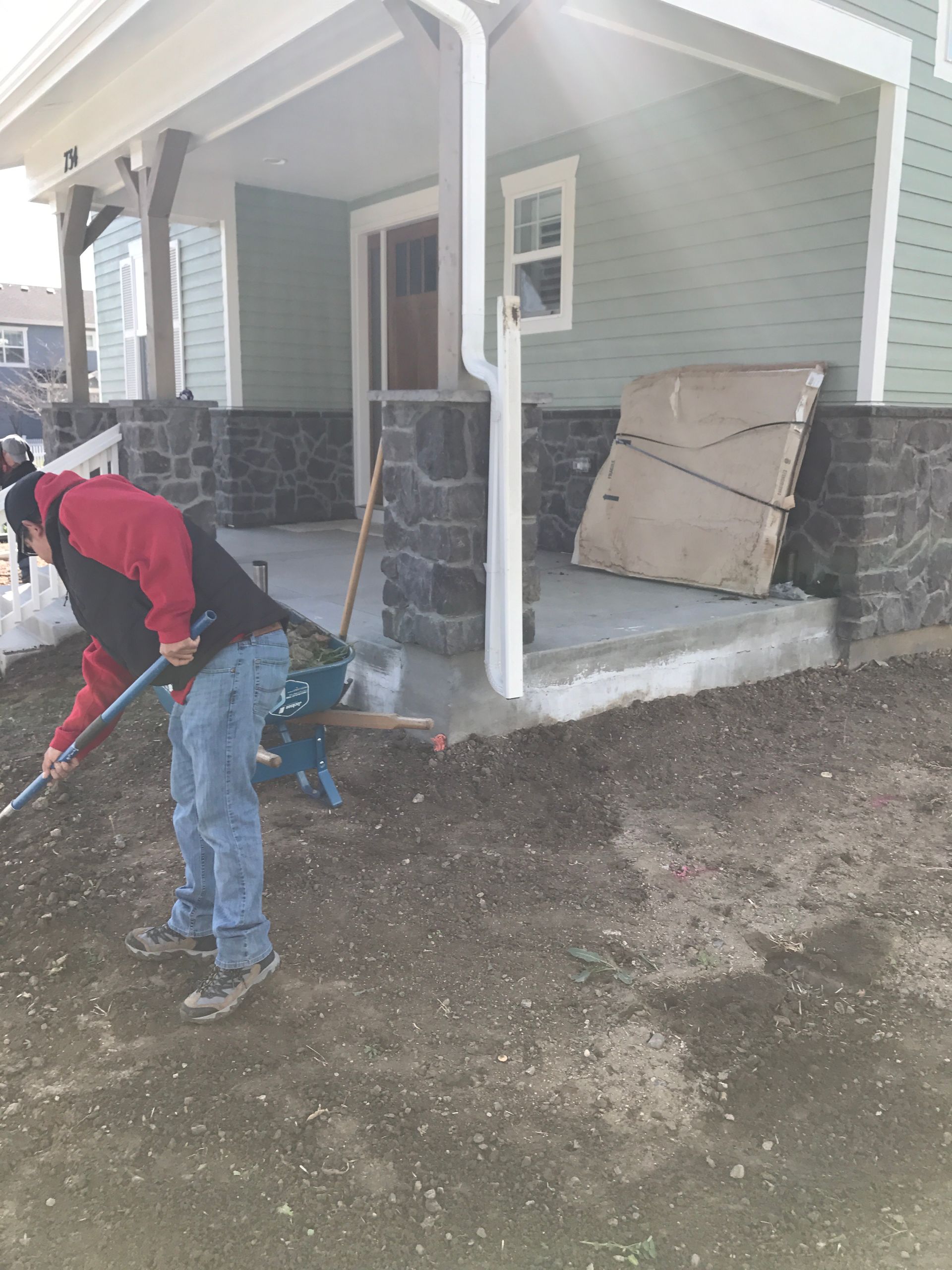 Two workers leveling ground near a house with a porch. One rakes, other appears to be preparing a cement slab.