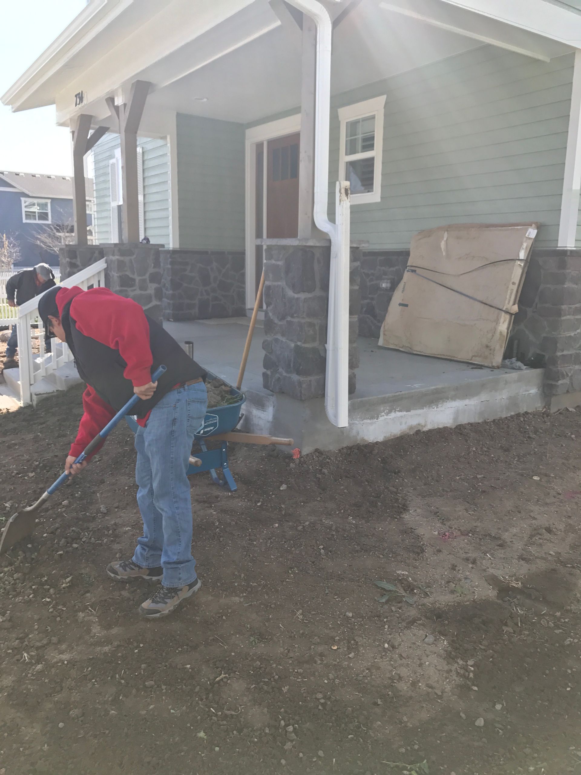 Man raking soil in front of a house with a porch, surrounded by building materials, cloudy sky.
