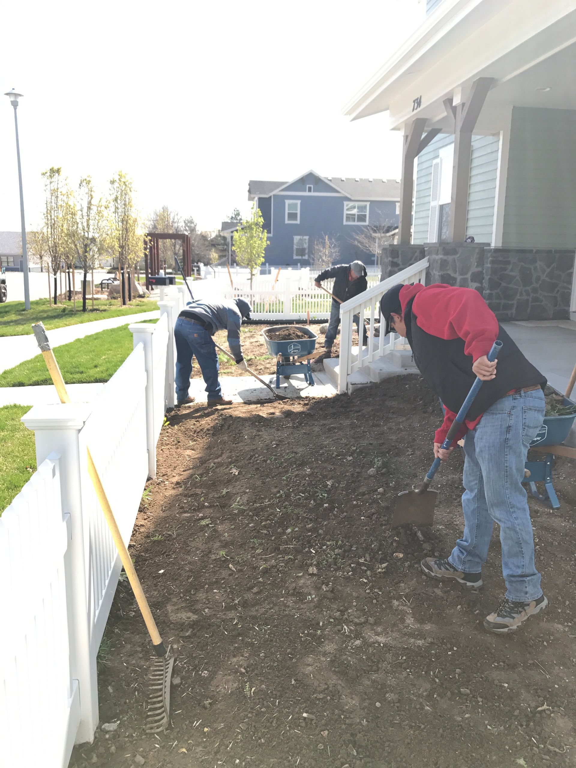 Three workers are landscaping a yard near a white picket fence, using tools in front of a house.