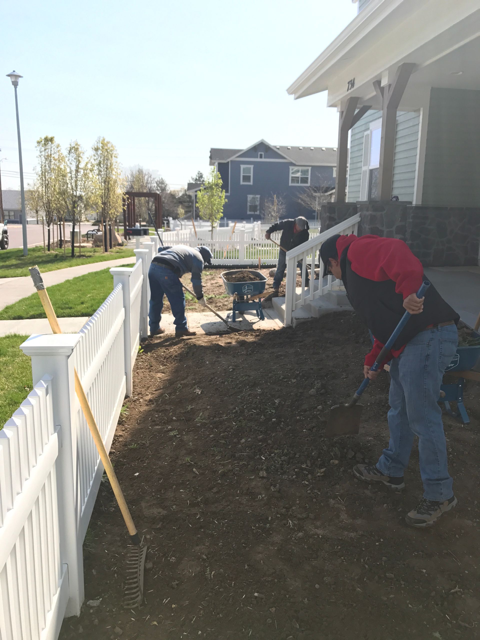 Men working in a yard near a white picket fence, prepping soil for planting.