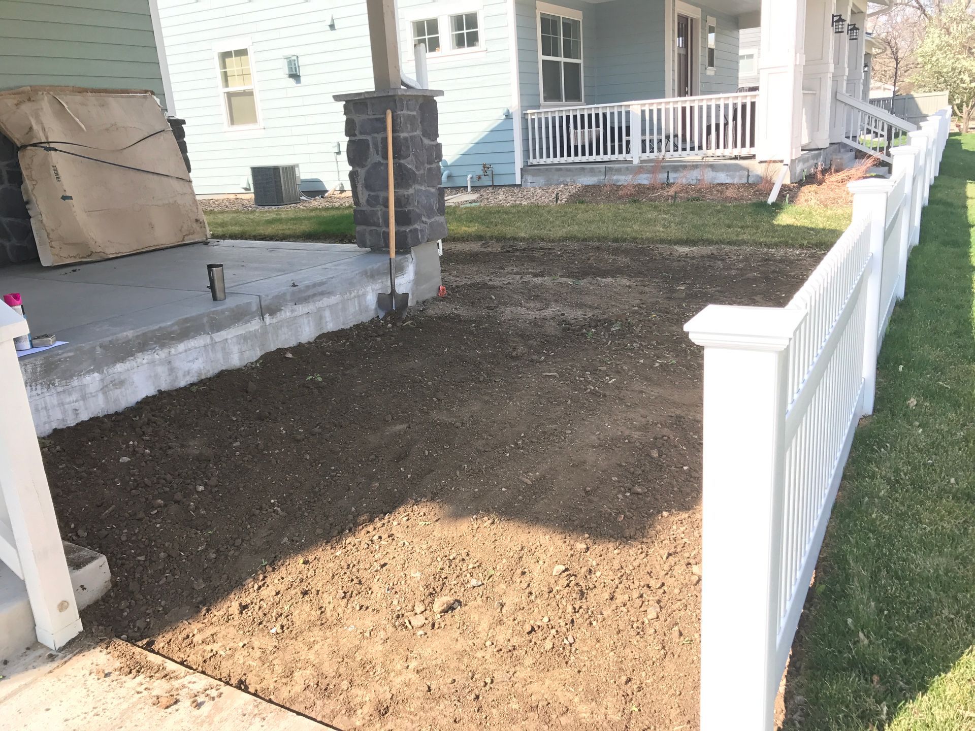 Newly laid soil in front of a house, beside a white picket fence, with porch in the background.