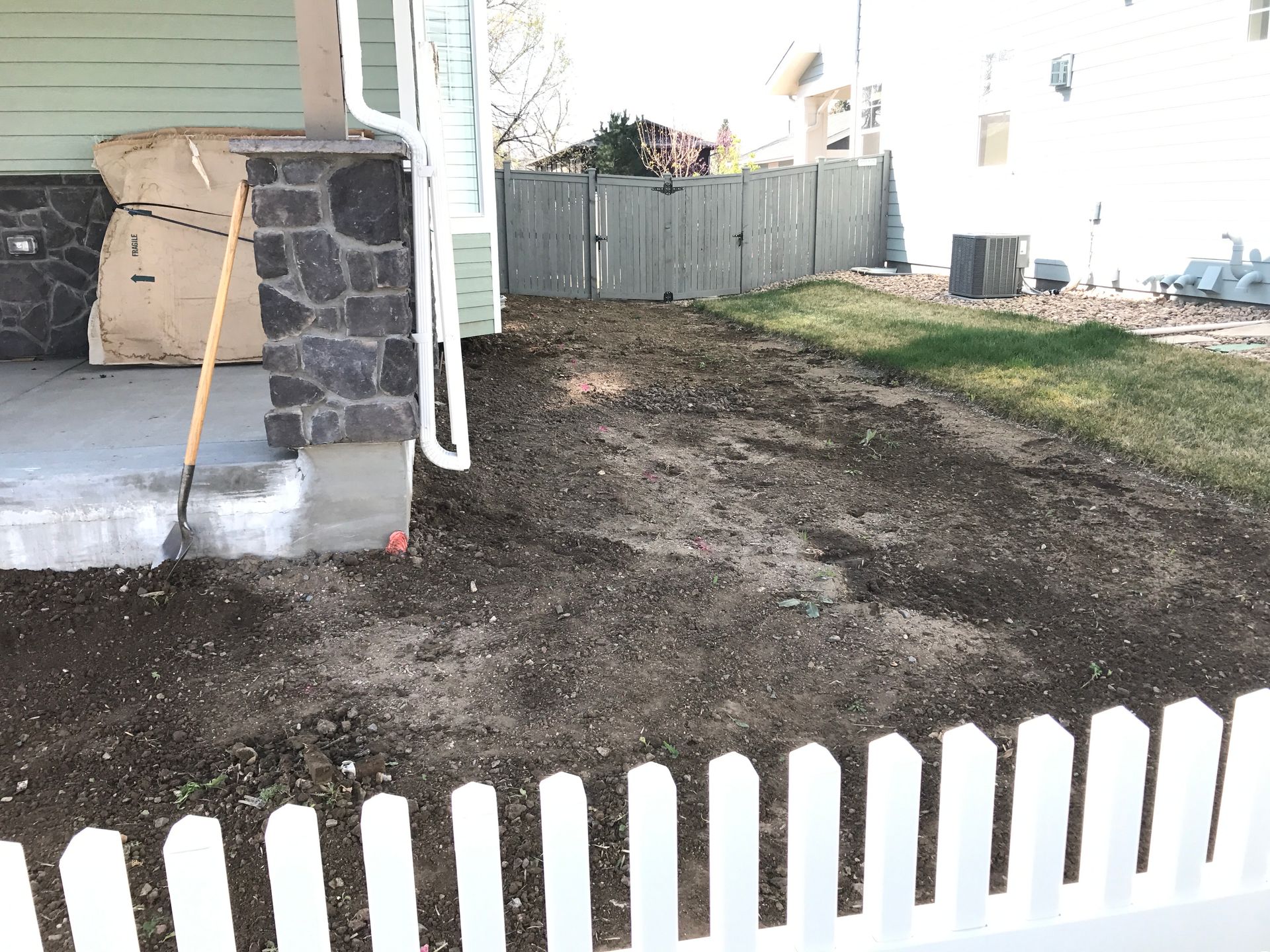 A front yard with a freshly tilled area next to a porch with a stone pillar, a picket fence is in the foreground.