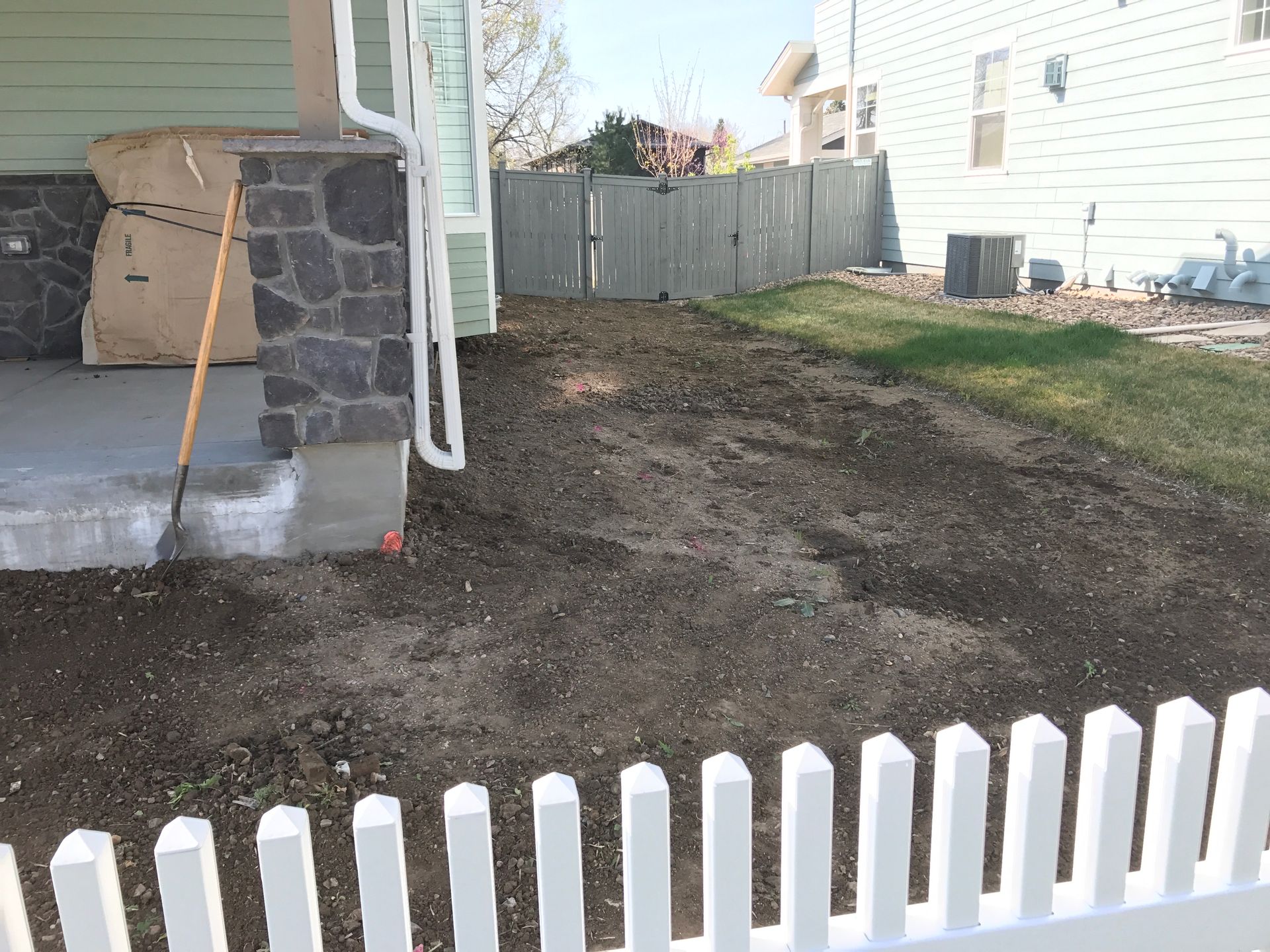 Dirt patch next to a house with a white picket fence in the foreground and a stone column.