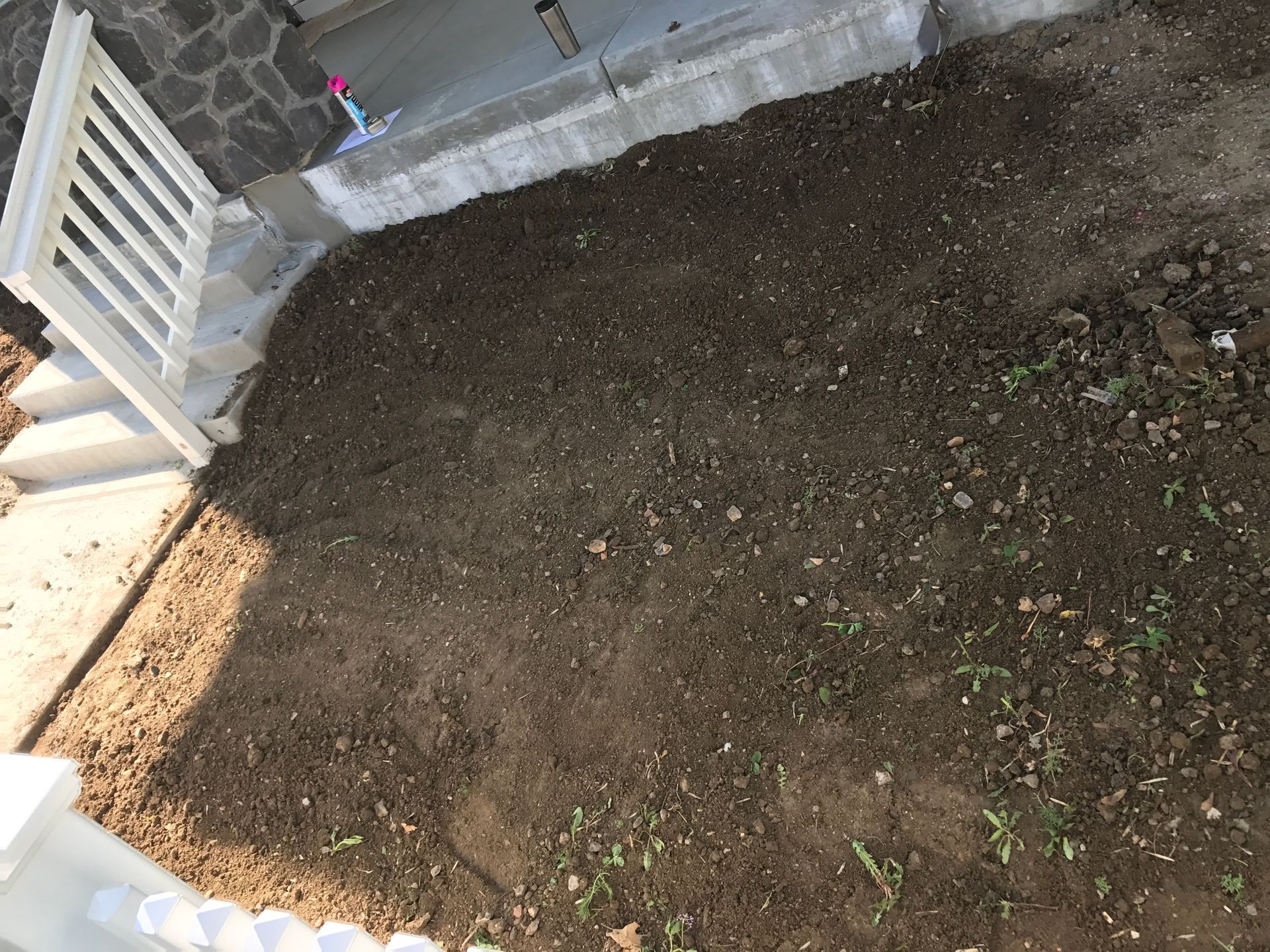 A freshly tilled, brown garden bed next to white stairs and railing, in front of a stone building.