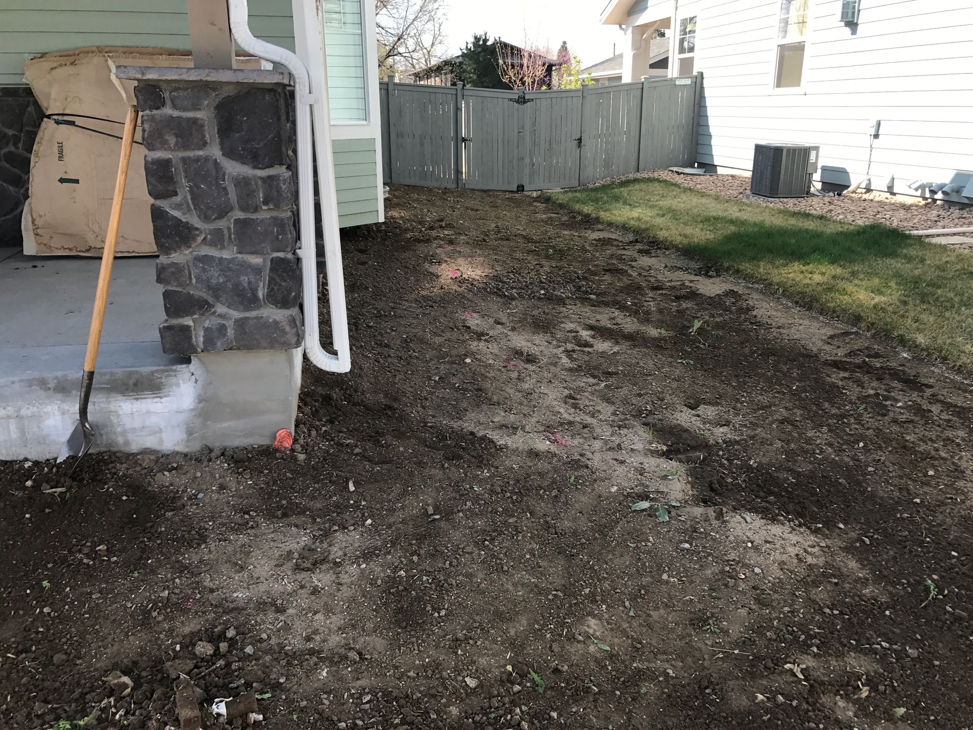 A dirt yard next to a house with a stone column, leading to a green fence and grass. A rake leans against the house.