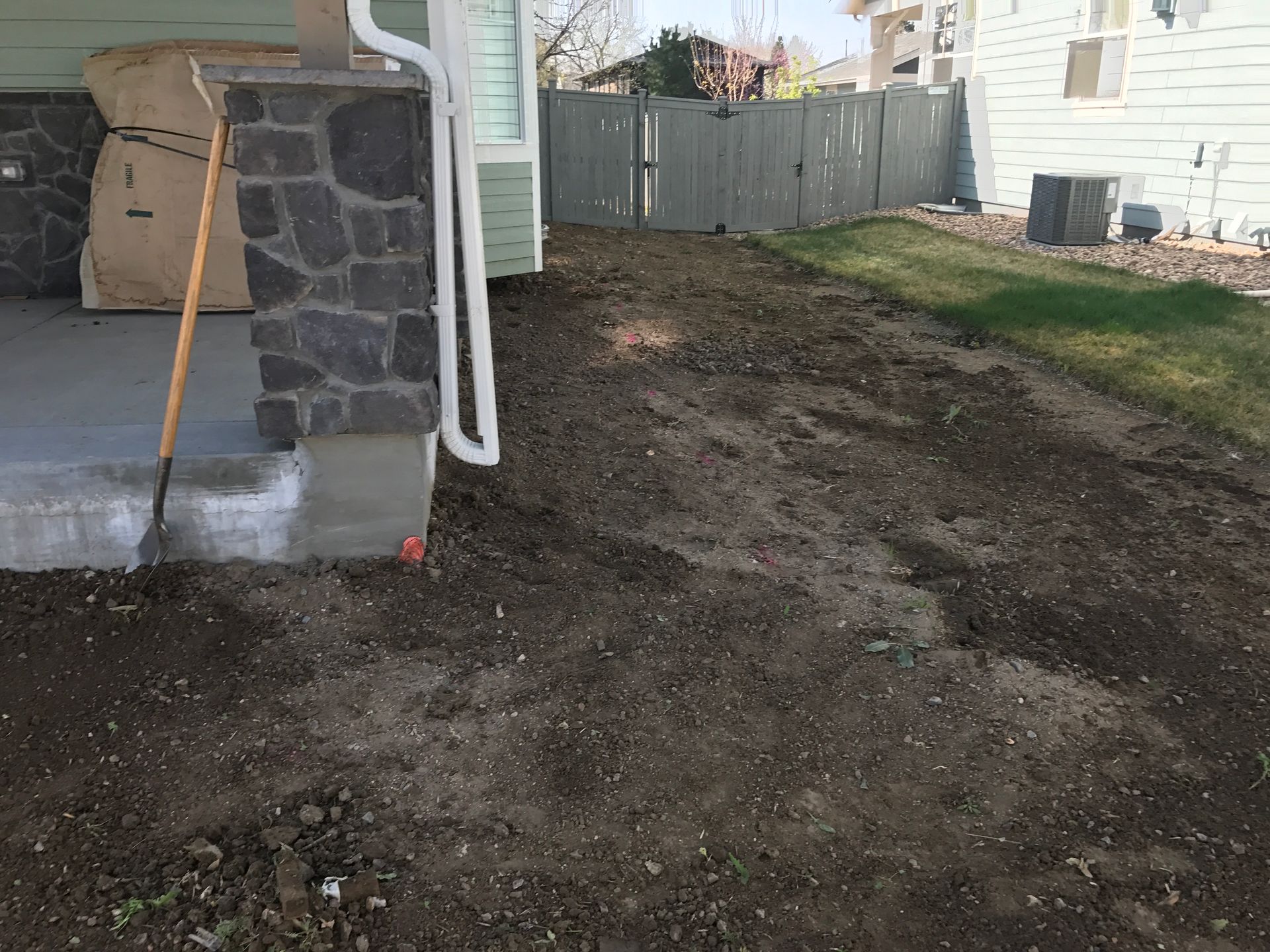 Dirt and grass yard next to a house with a concrete porch and stone pillar. A shovel leans against the porch.