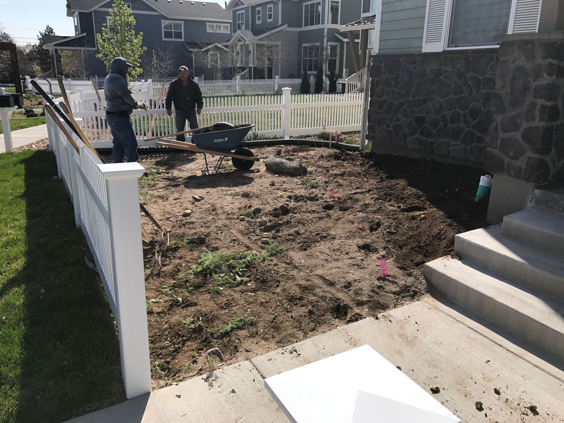 Two men working on a dirt patch in front yard, with a white picket fence and house in the background.