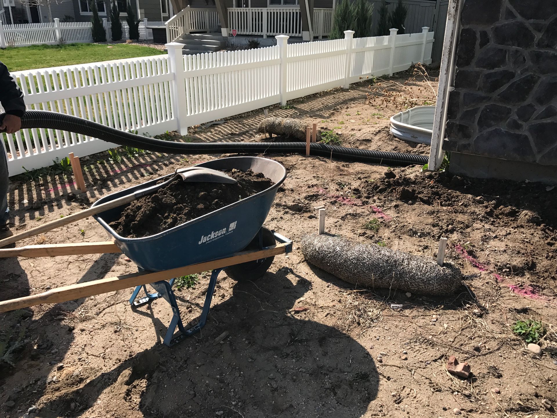 A person filling a wheelbarrow with dirt near a white picket fence, a house, and drainage pipes.