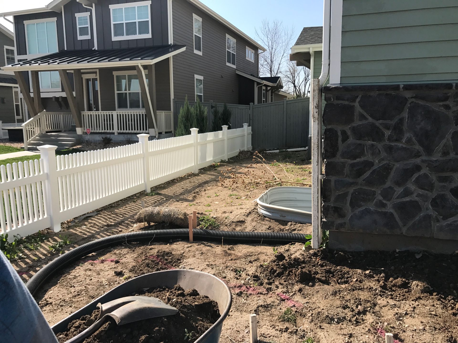 A backyard with a white picket fence, dirt, black drainage pipe, and a wheelbarrow.