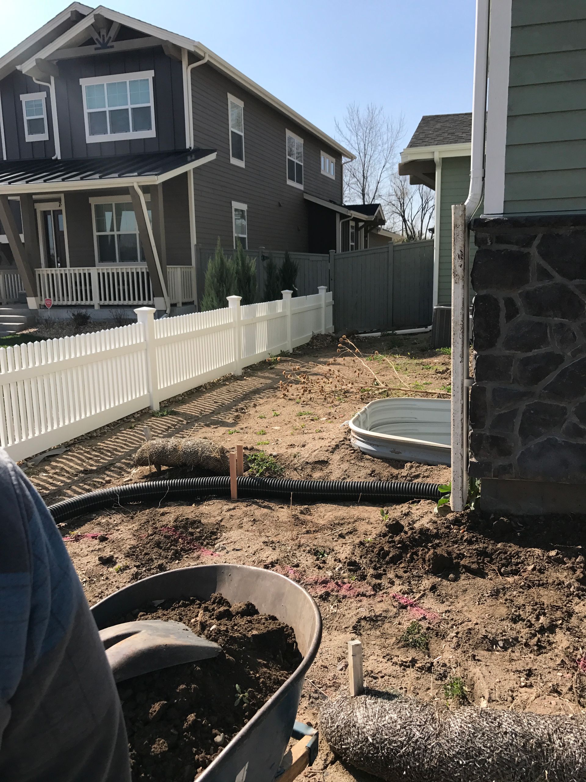 Backyard with white picket fence, dirt, wheelbarrow, two-story houses in background.