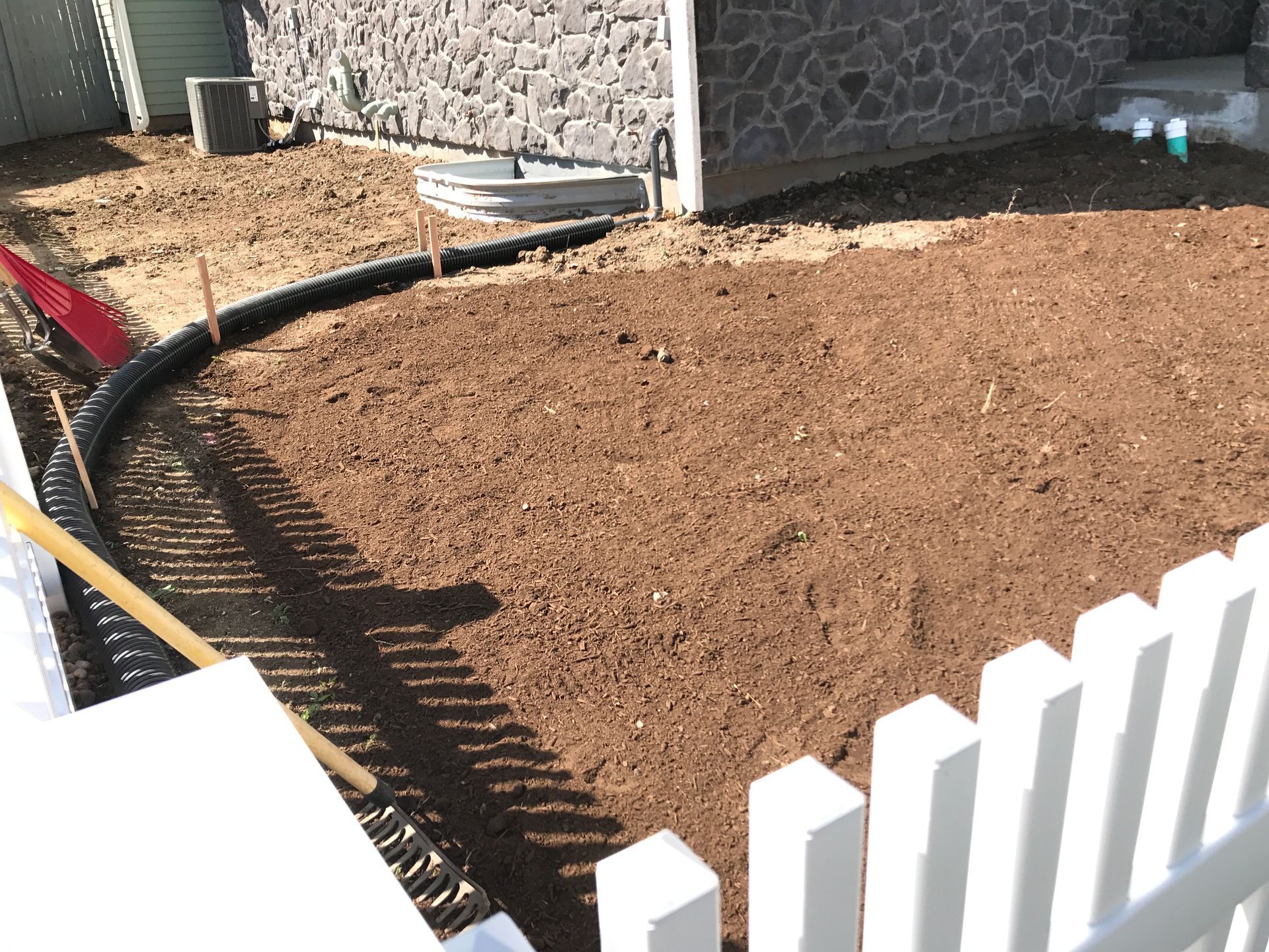 Prepared soil bed with curved edging, bordered by a white picket fence, near a house with stone siding.