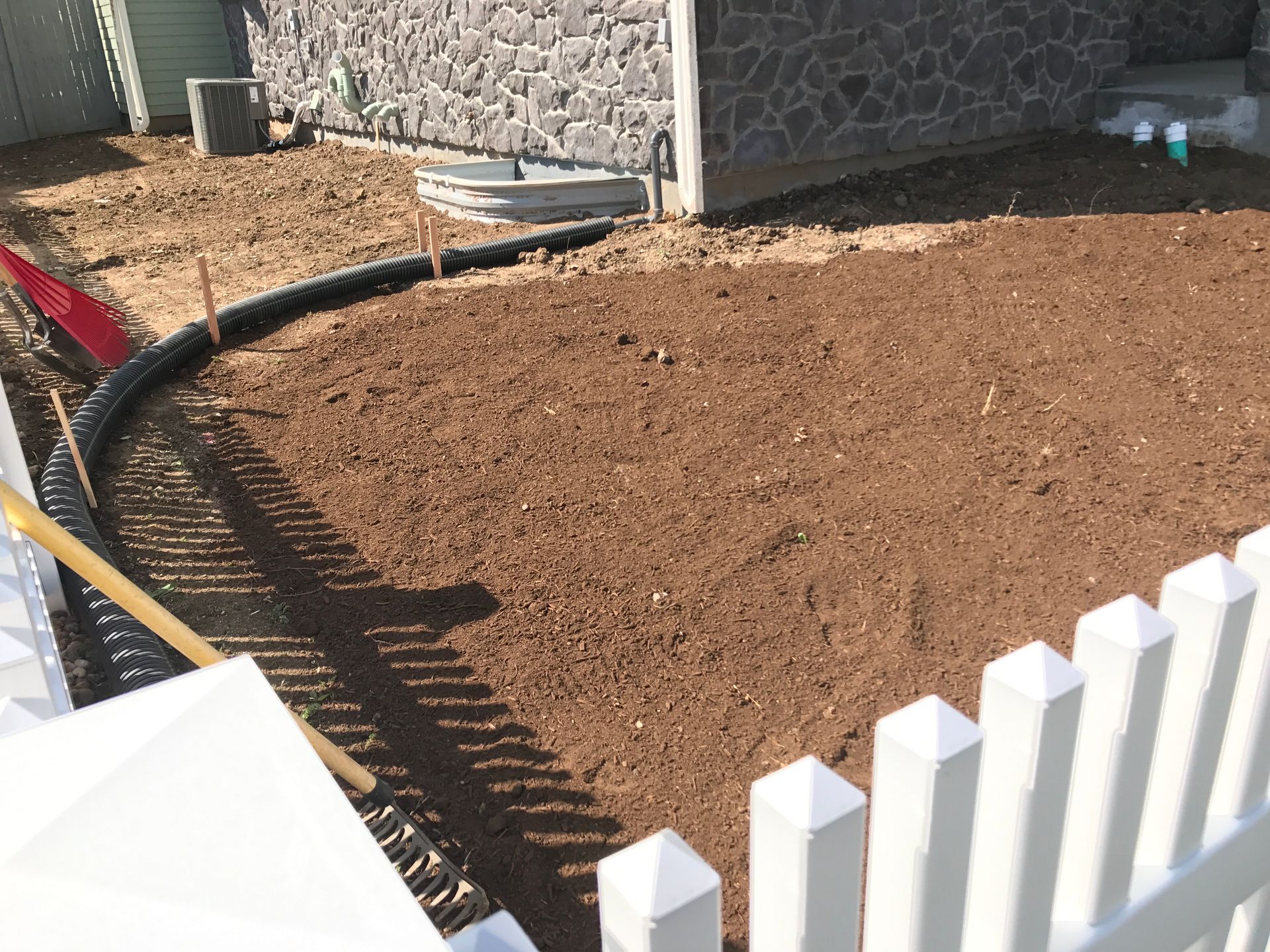 A prepared garden bed of brown soil next to a house with a white picket fence.