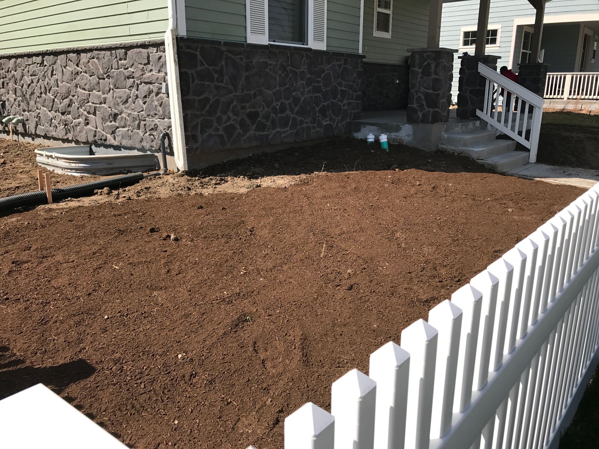 Front yard with fresh soil, white picket fence, and house with stone foundation.