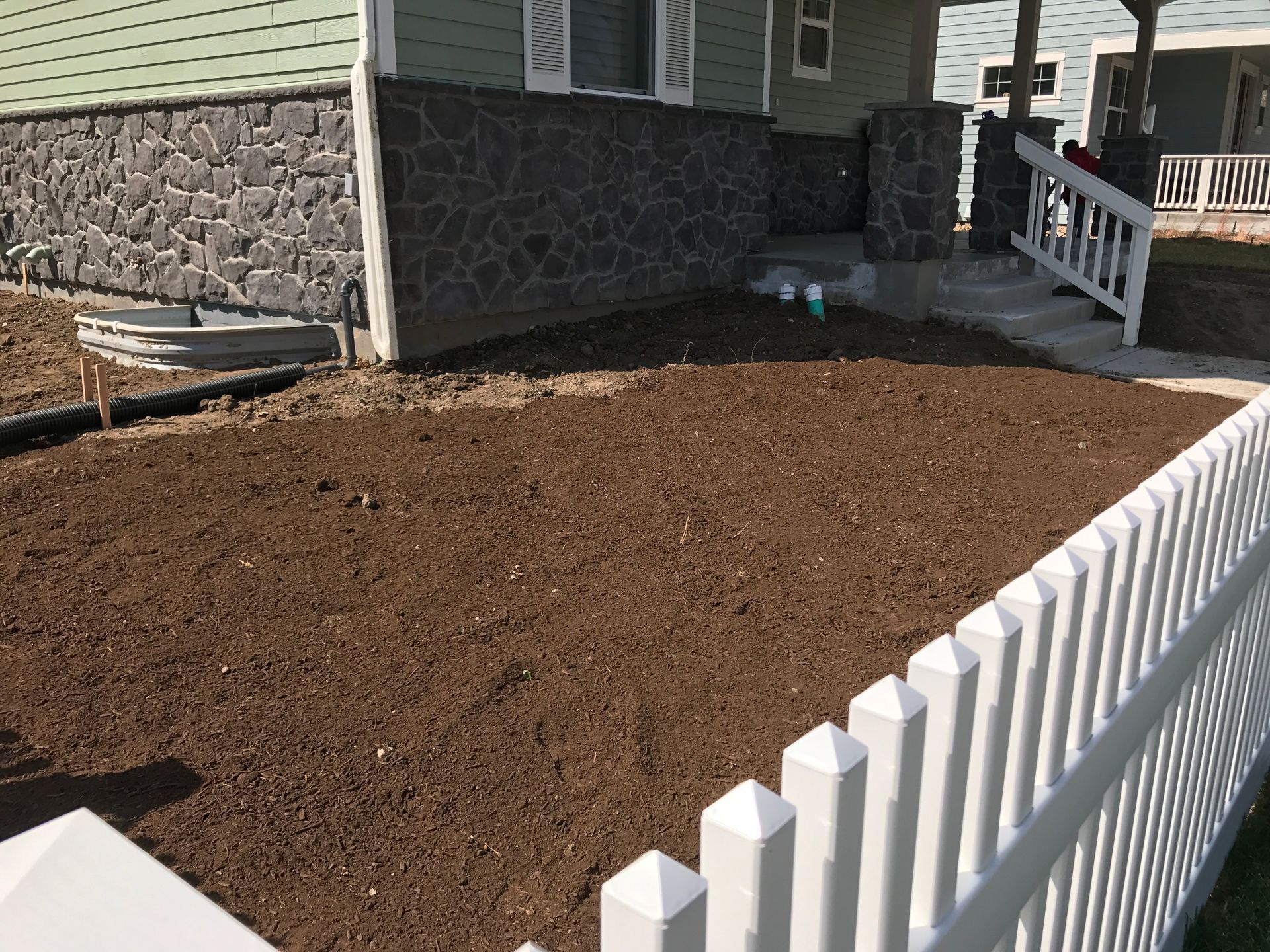 A yard with freshly turned brown soil in front of a house with a white picket fence.