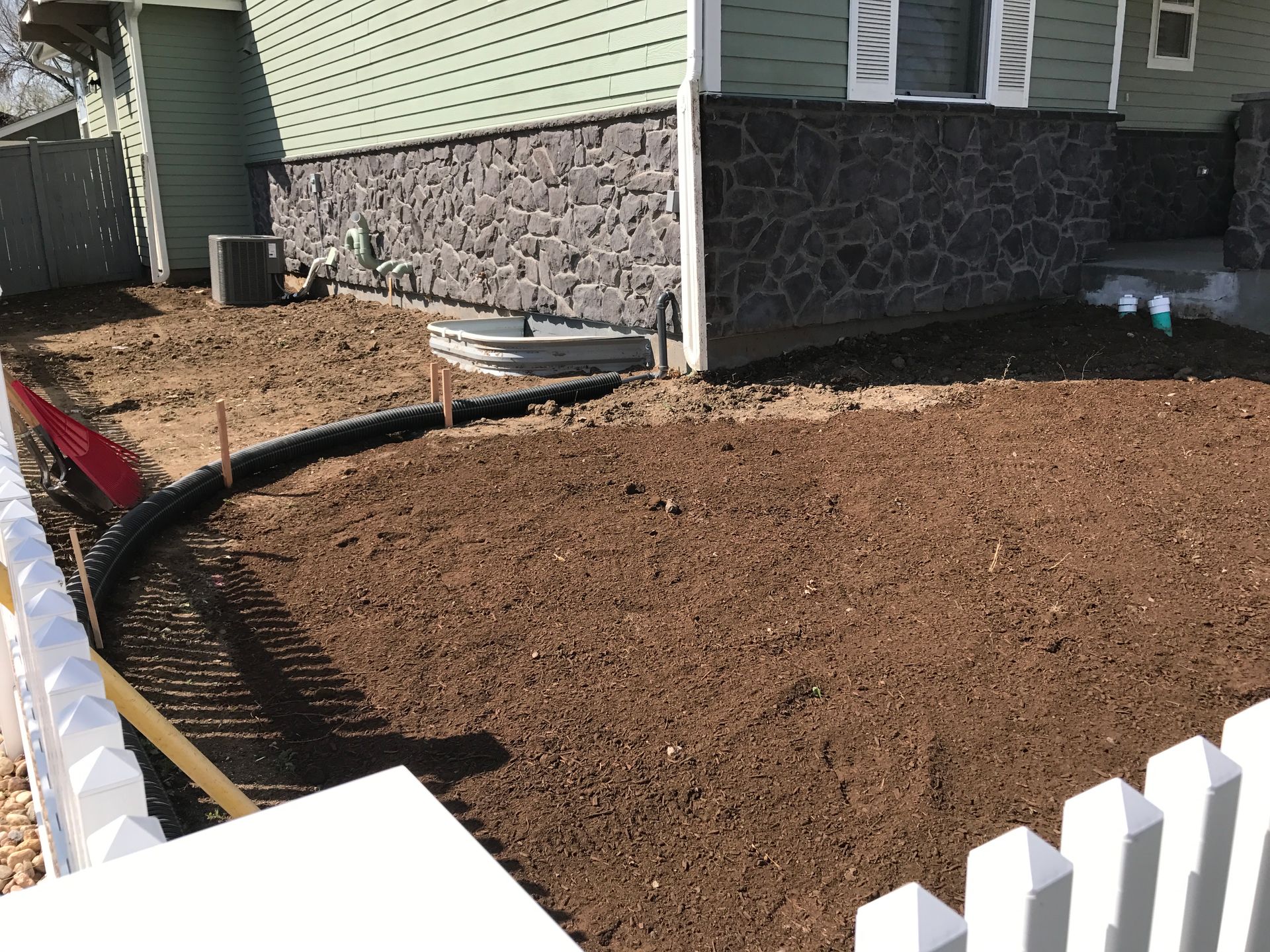 A front yard with recently tilled dark soil, a black drainage pipe, and a white picket fence bordering the area.