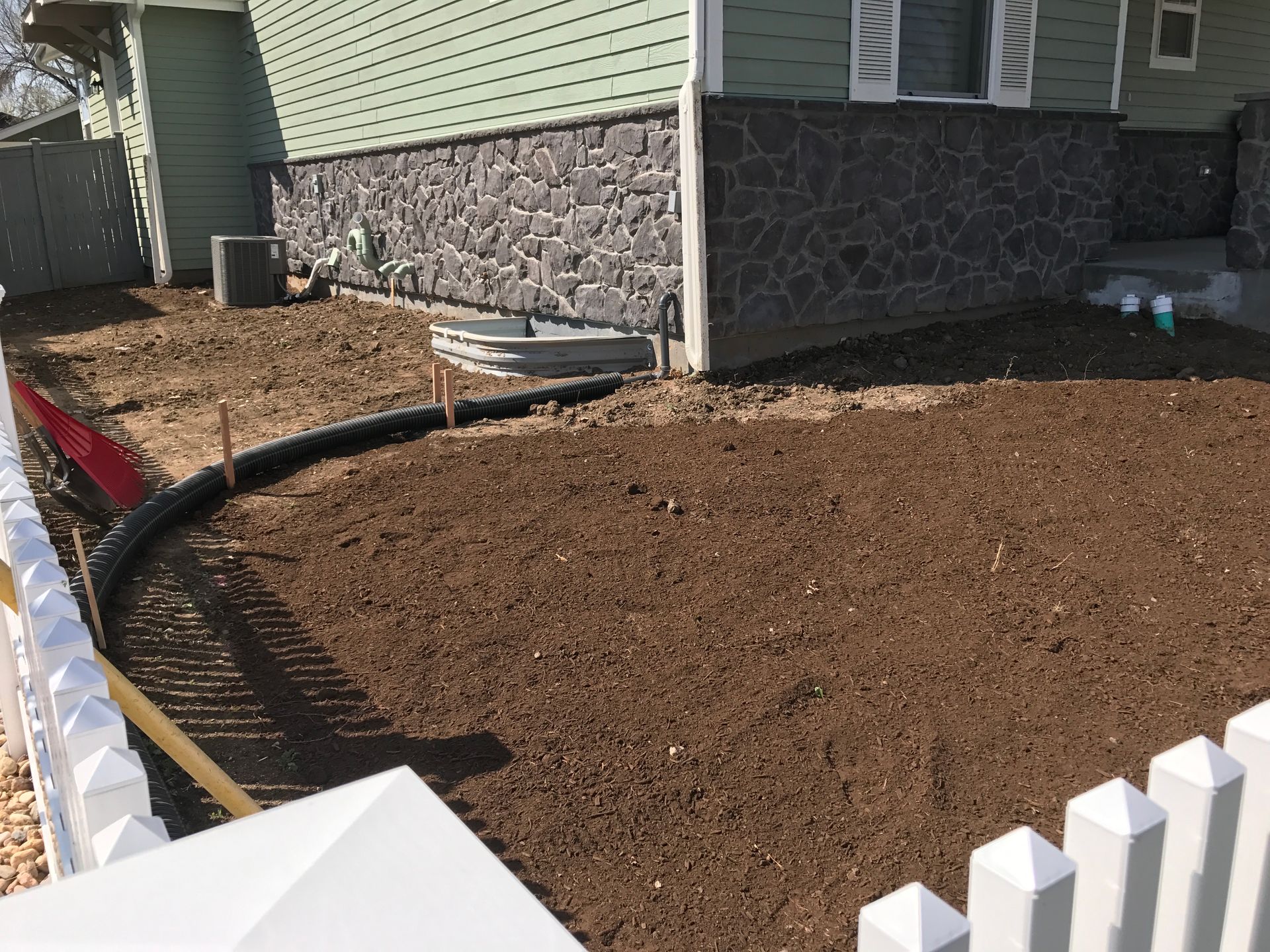 Freshly tilled brown soil in front of a house, bordered by a black curve and a white picket fence.