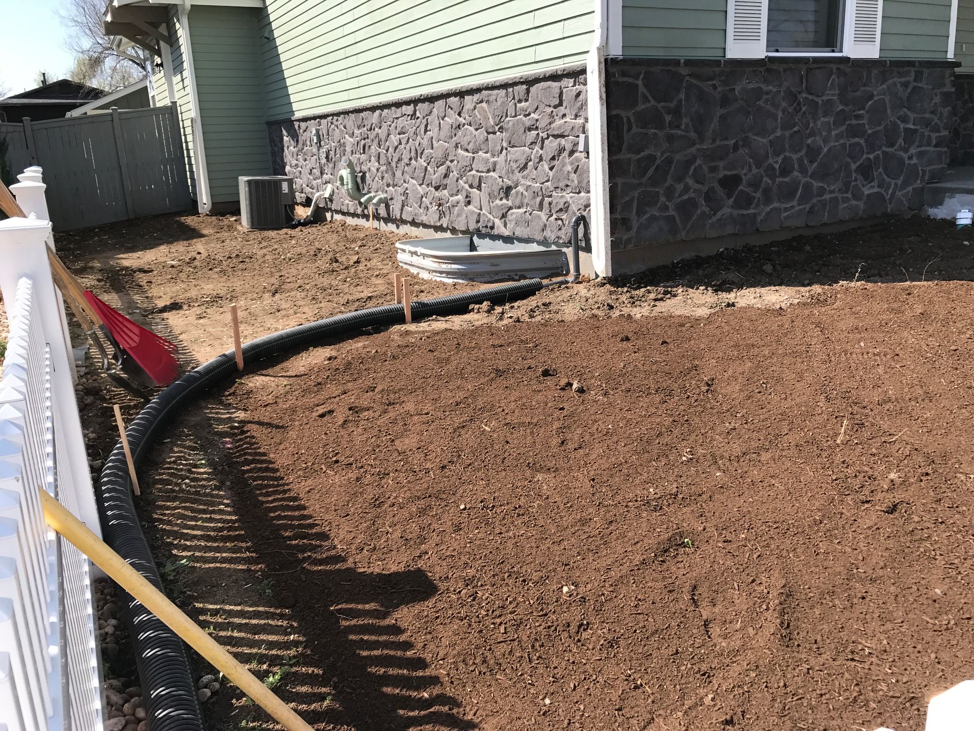 A house with a black stone facade, newly tilled dirt, and a black drainage pipe next to a white fence.