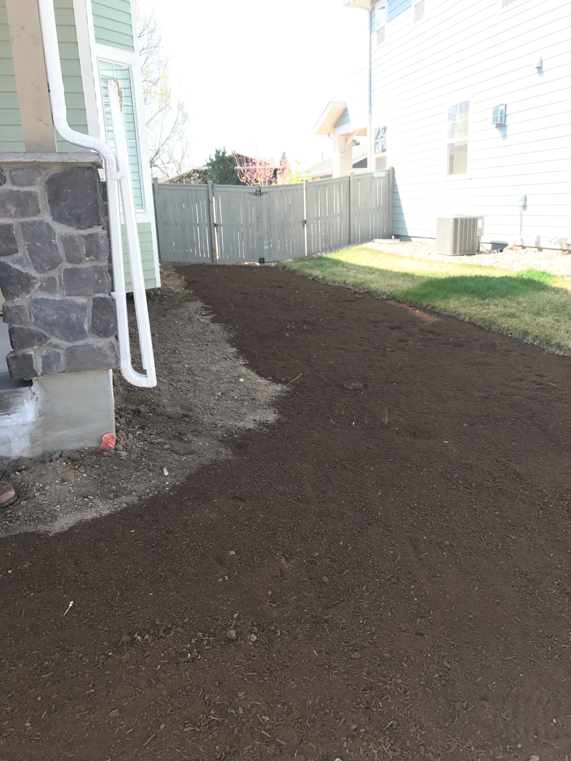 Dark brown soil pathway beside a house, leading to a wooden fence. White drainpipe on the left.