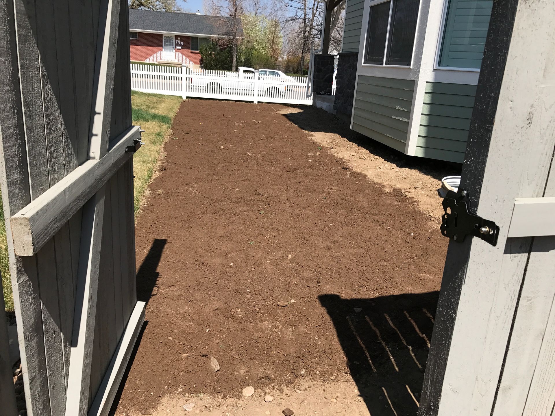 Open wooden gate leading to a freshly tilled garden bed between a house and lawn.