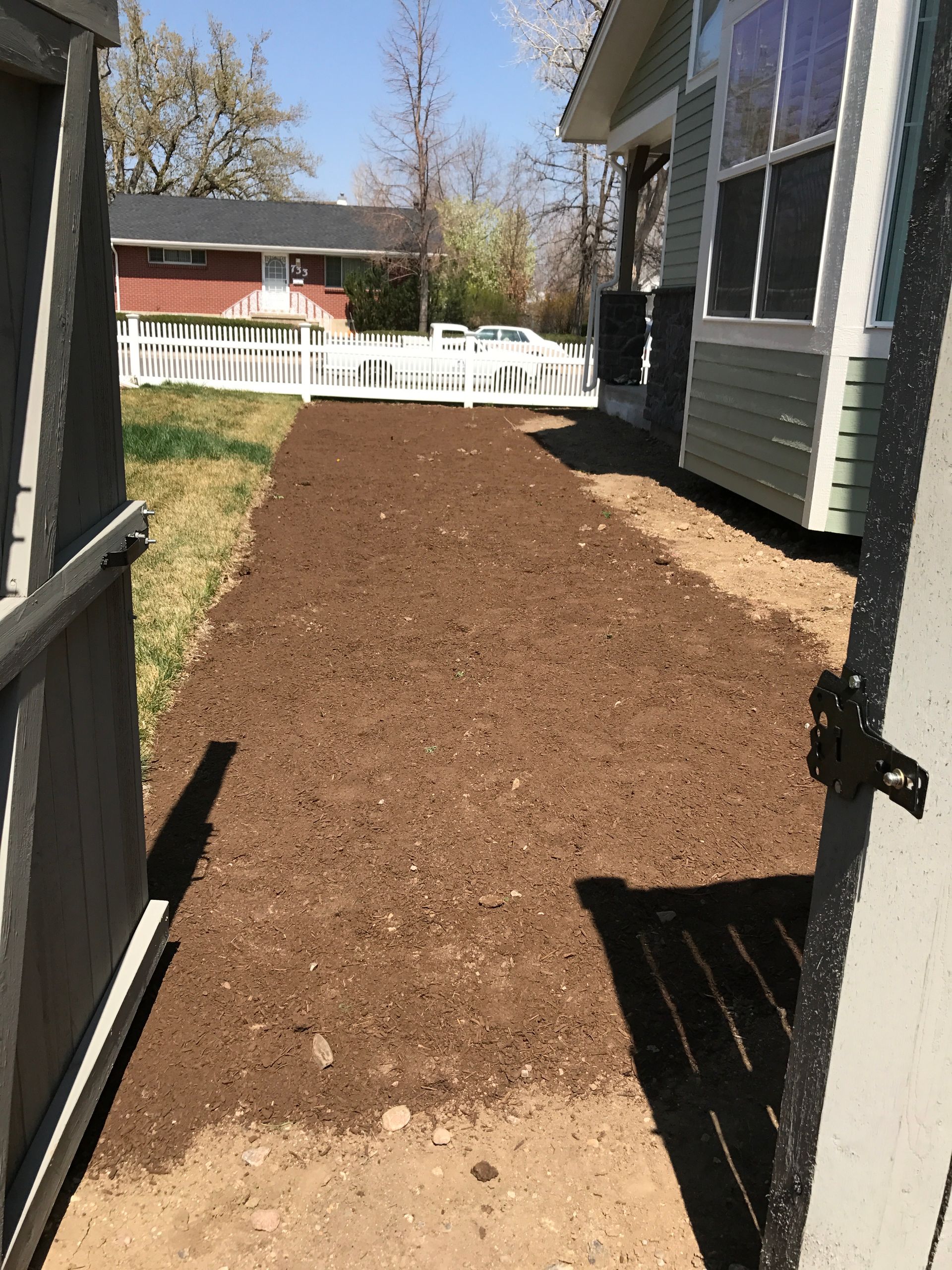 Gravel path between house and fence. Dark brown earth is tilled, ready for planting. Gate on left is open.