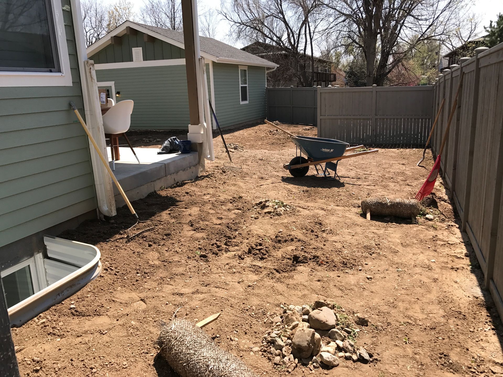 Backyard dirt area; wheelbarrow, fence, small building, and partially visible house on a sunny day.
