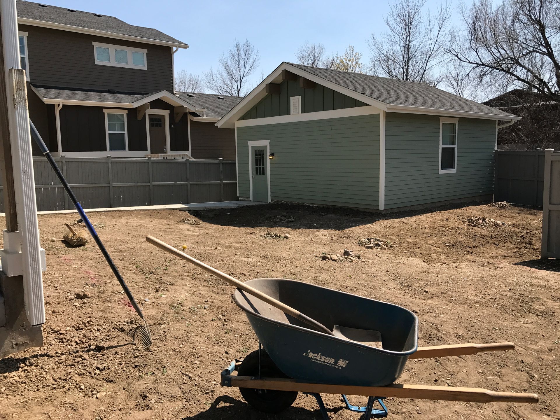 Backyard with a wheelbarrow, rake, and shed. Two-story house in background.