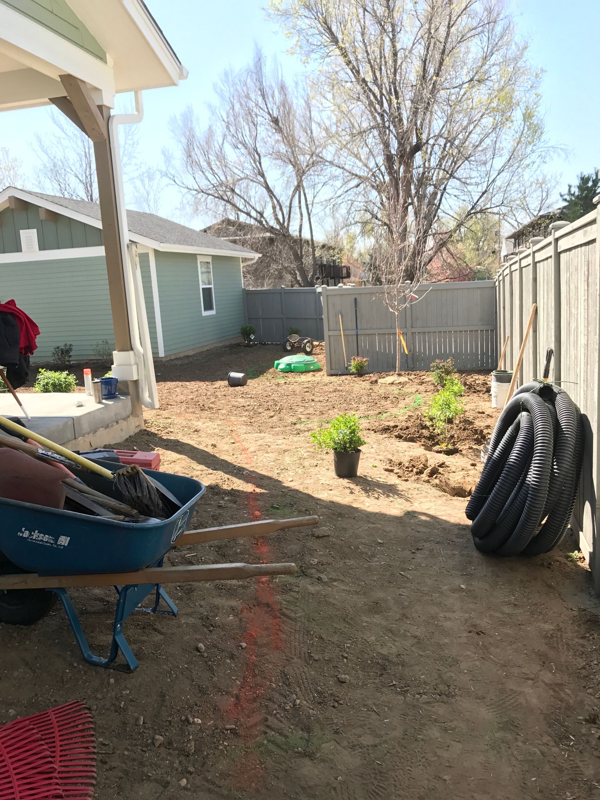 Backyard under construction with wheelbarrow, tools, and dirt. Green house and fence in background.