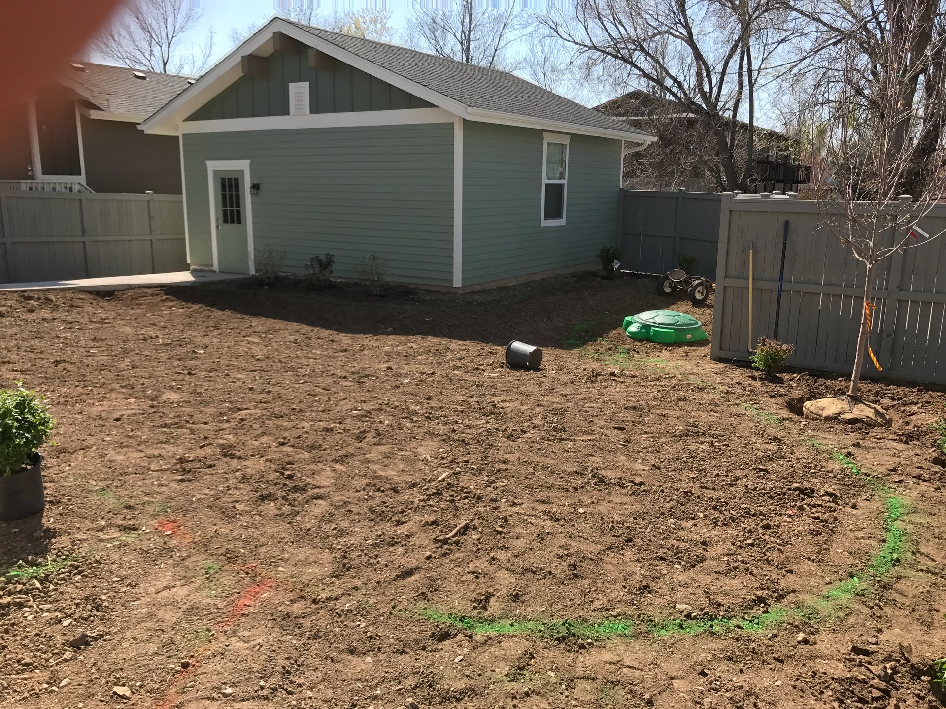 Backyard with a green shed and a dirt ground, surrounded by a gray fence, under a bright sky.
