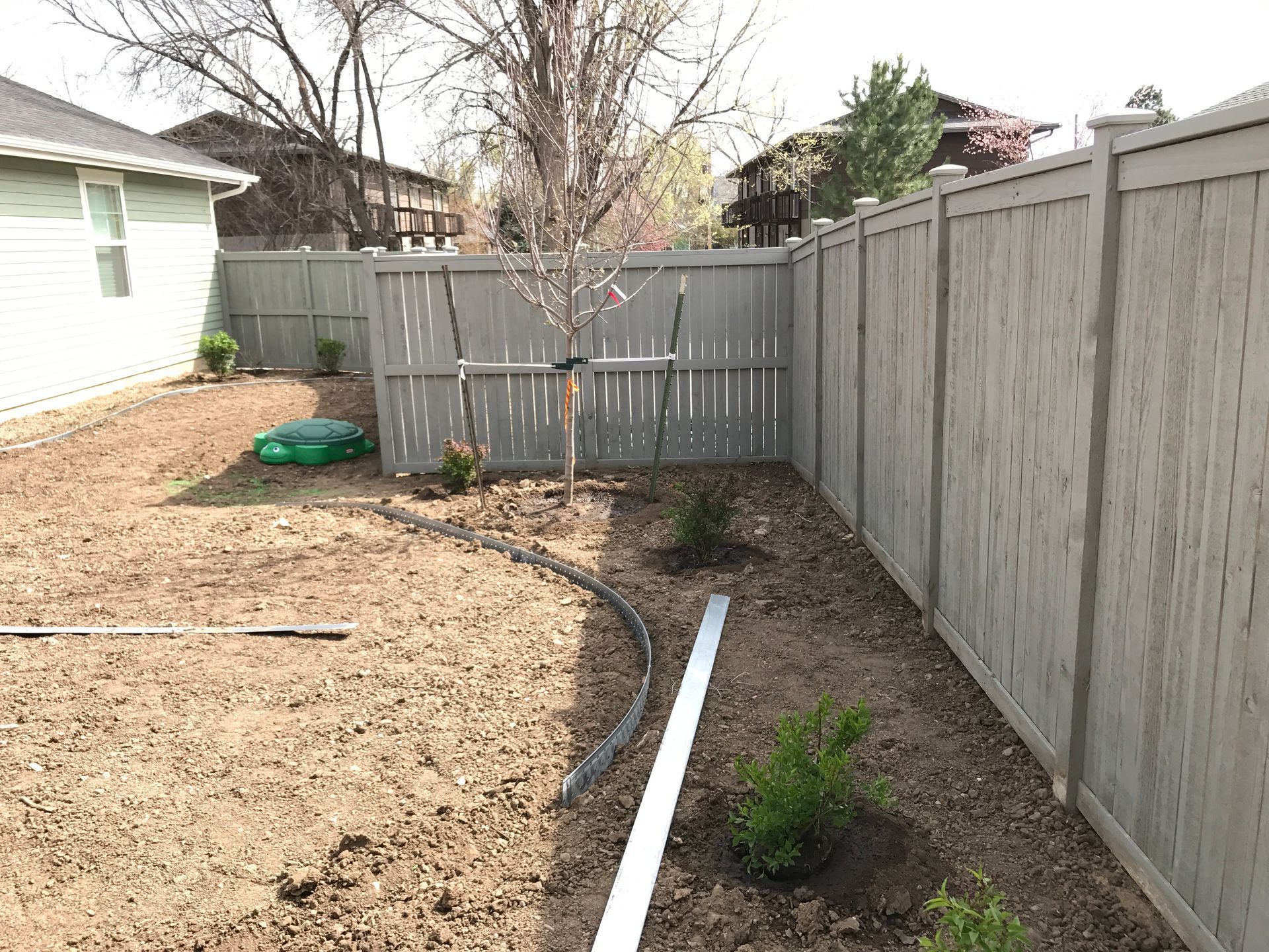 Backyard with gray fence, bare tree, and brown dirt.