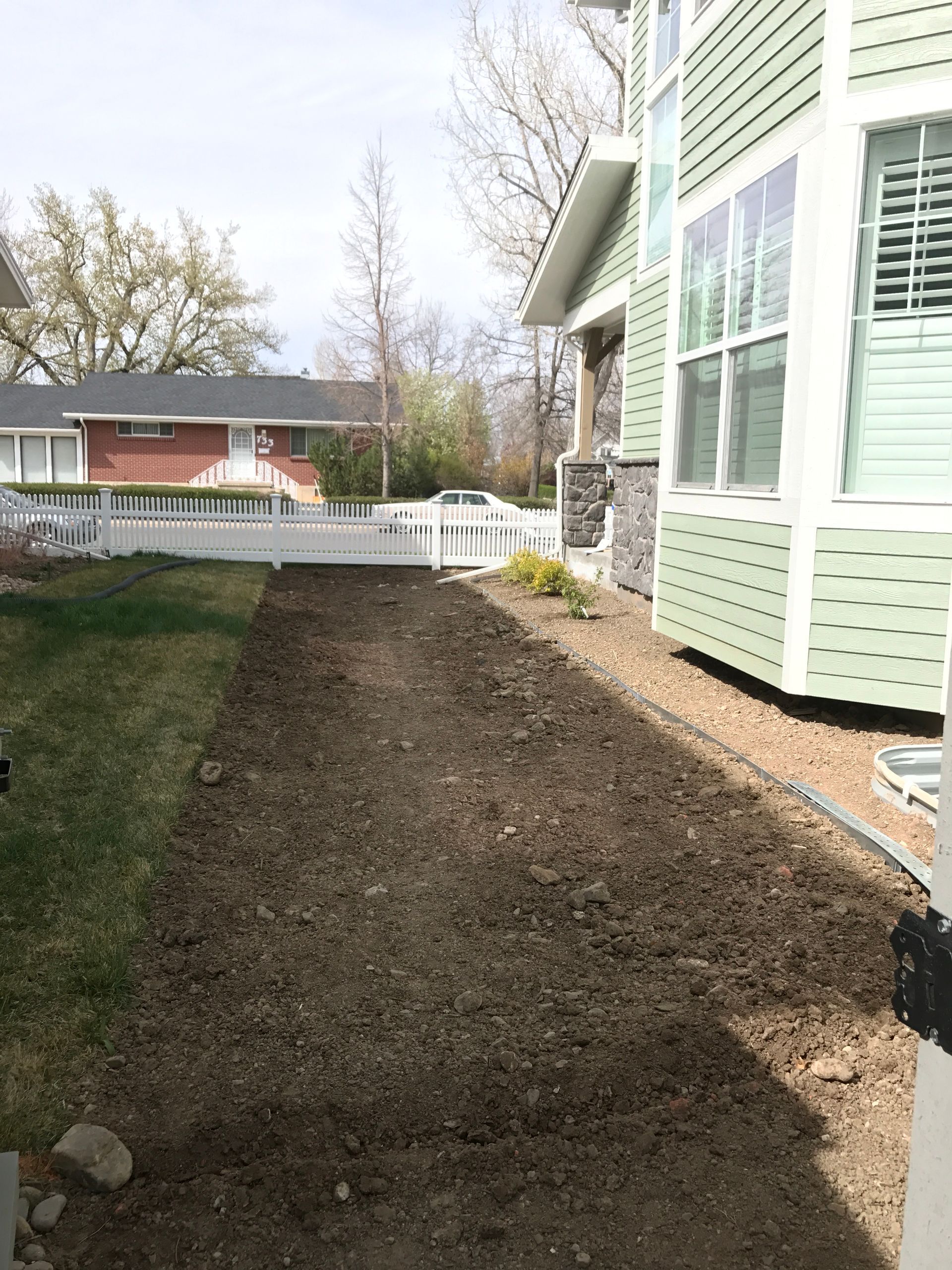 A strip of freshly tilled brown soil next to a light green house and white picket fence in a yard.