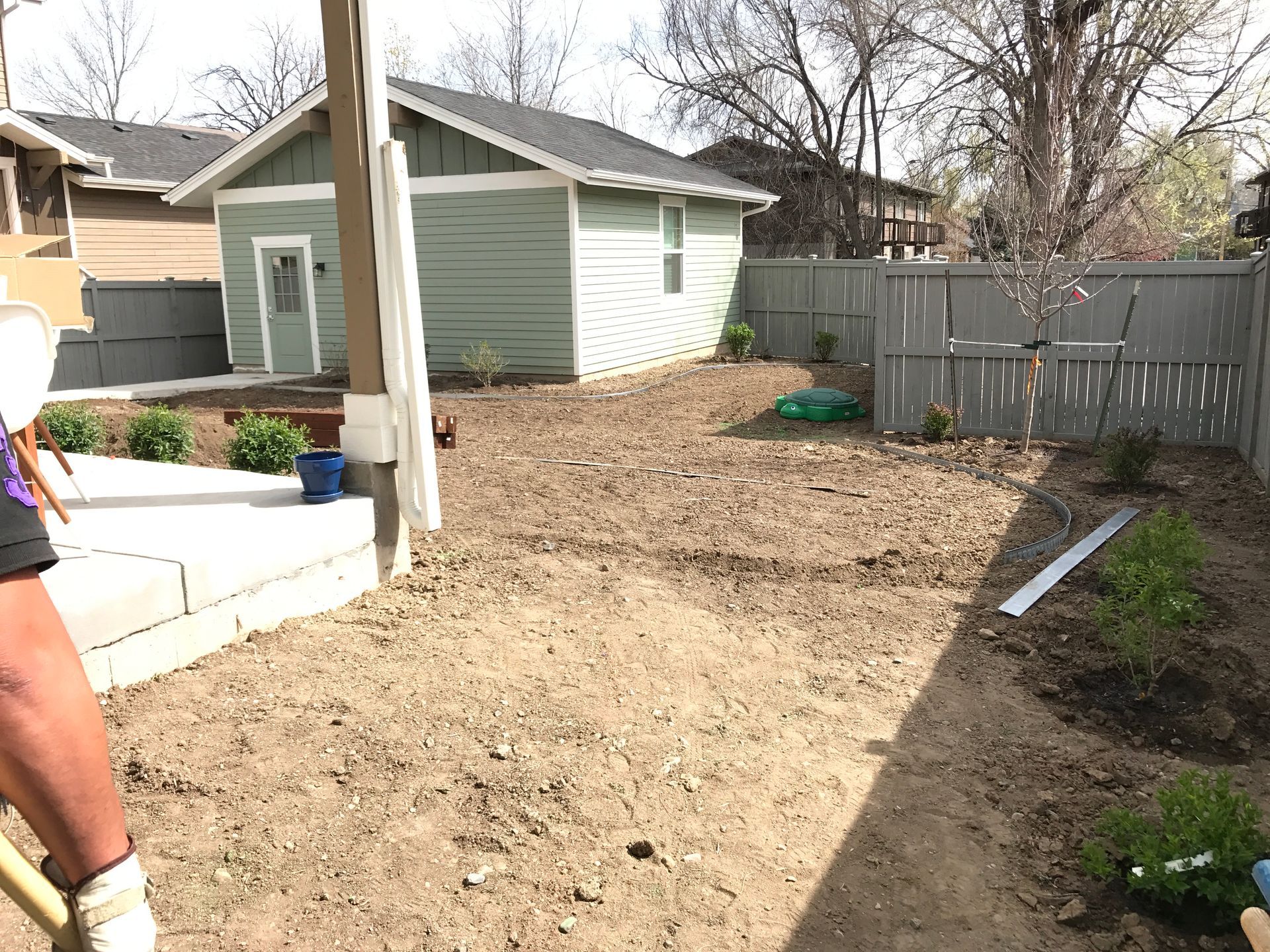 Backyard with dirt patch, small plants, green shed, gray fence, person holding a shovel.