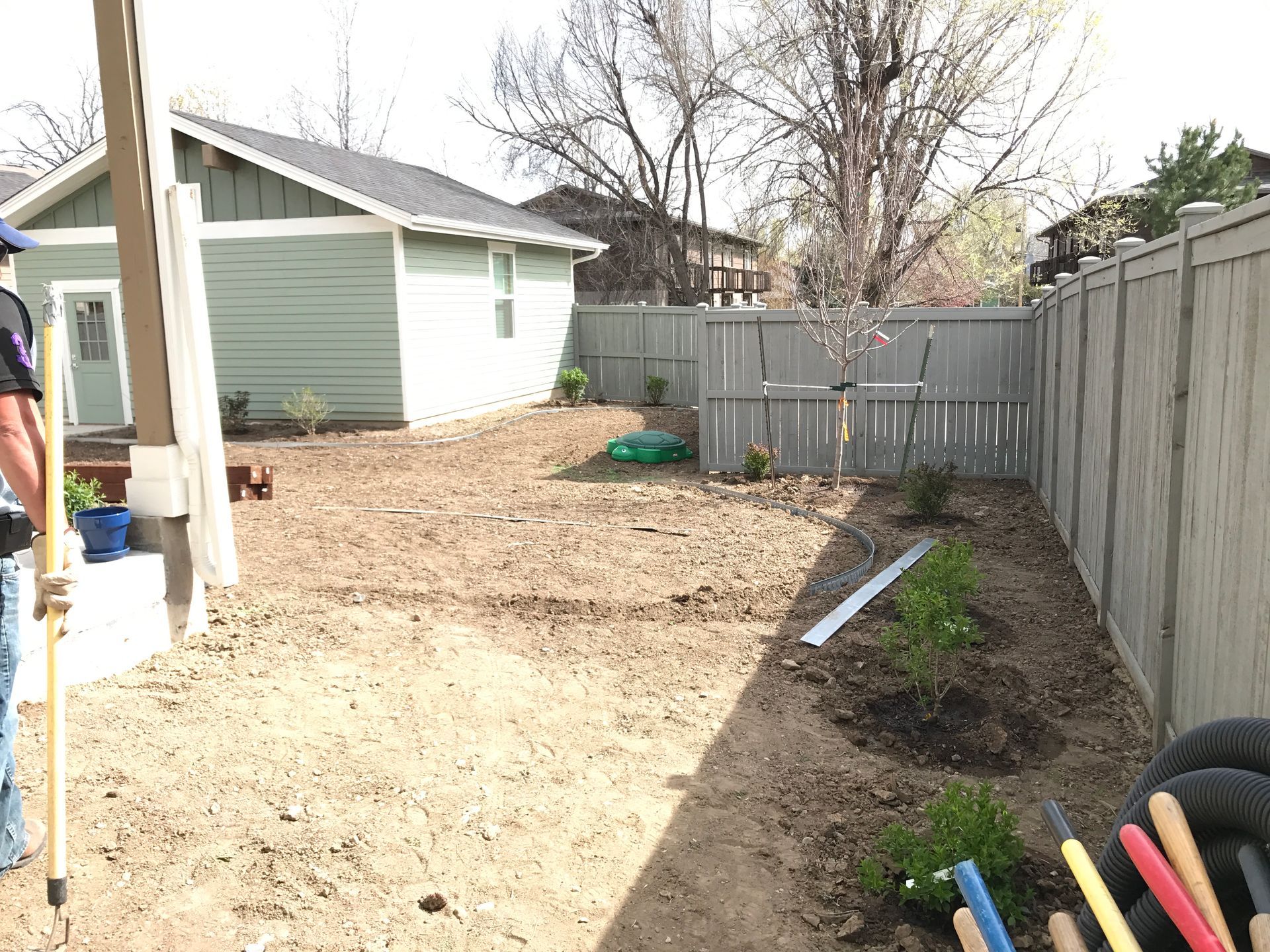 Backyard with dirt, fencing, a small green building, and someone holding a tool.