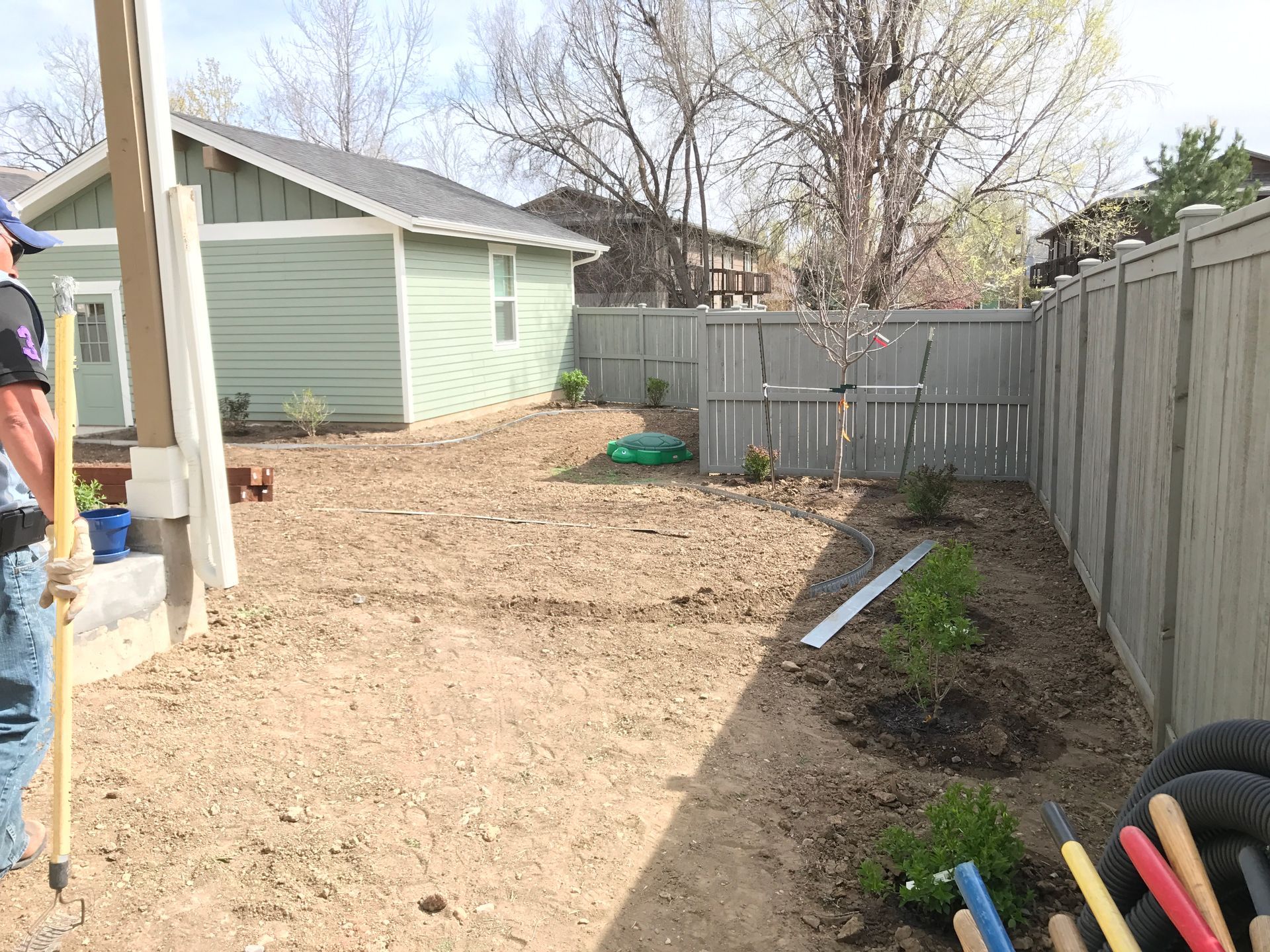 Backyard with dirt ground, small building, gray fence, and a person holding a pole.