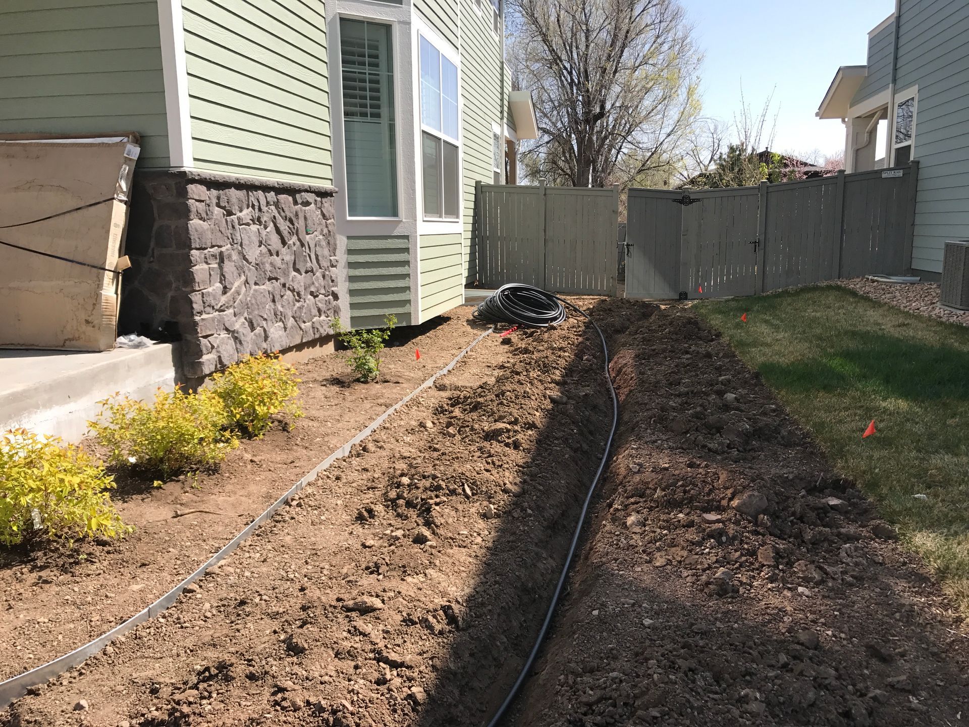 A trench dug along a building's foundation, with black tubing inside, and small bushes.