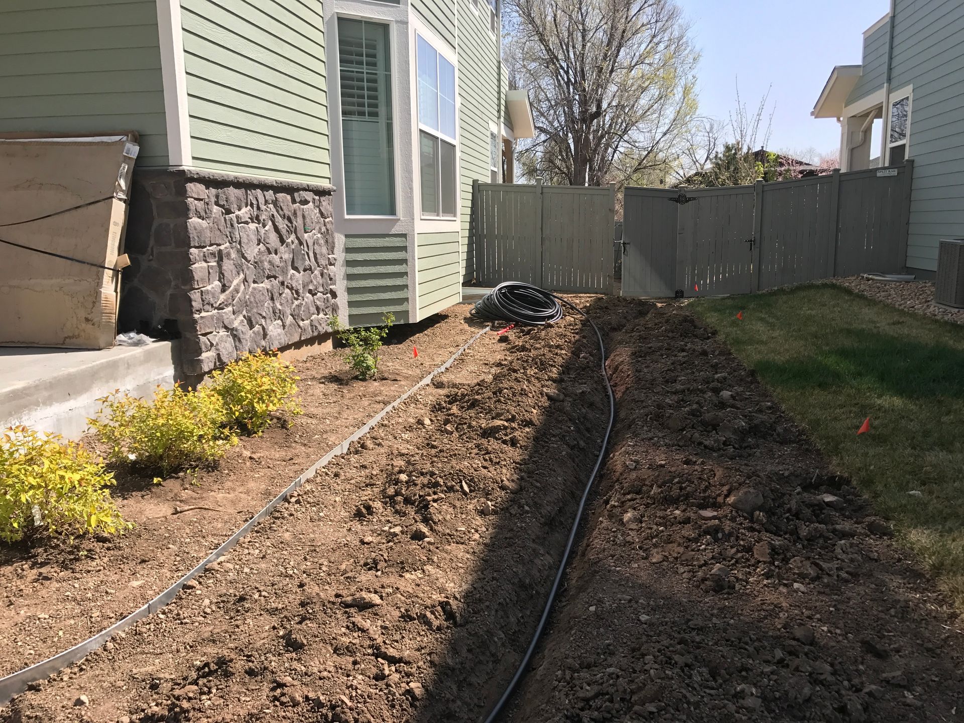 Black tubing in a dirt trench, along a house with a stone and green siding, and a wooden fence.