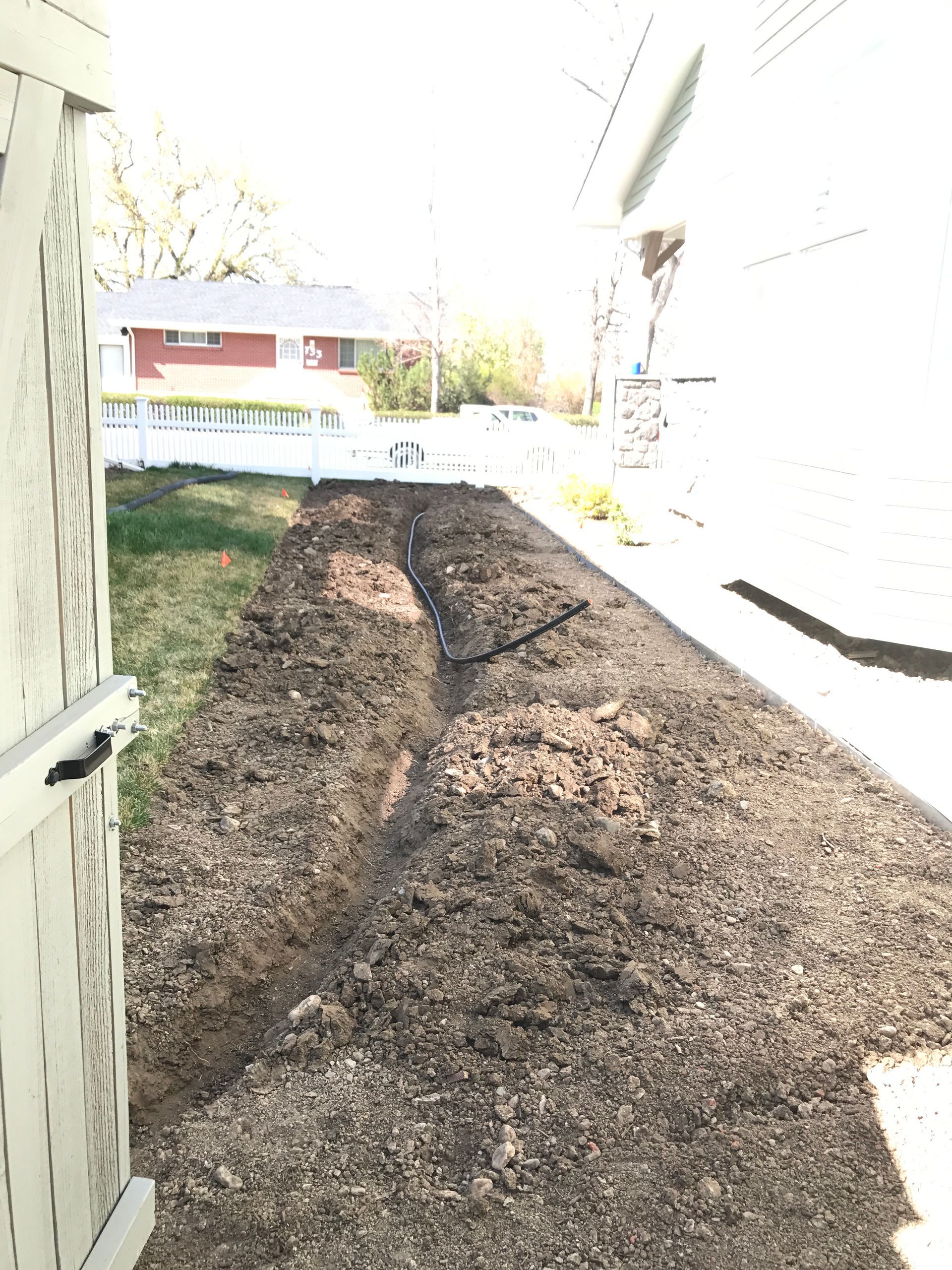 A long garden bed of dark soil next to a house and fence, ready for planting.