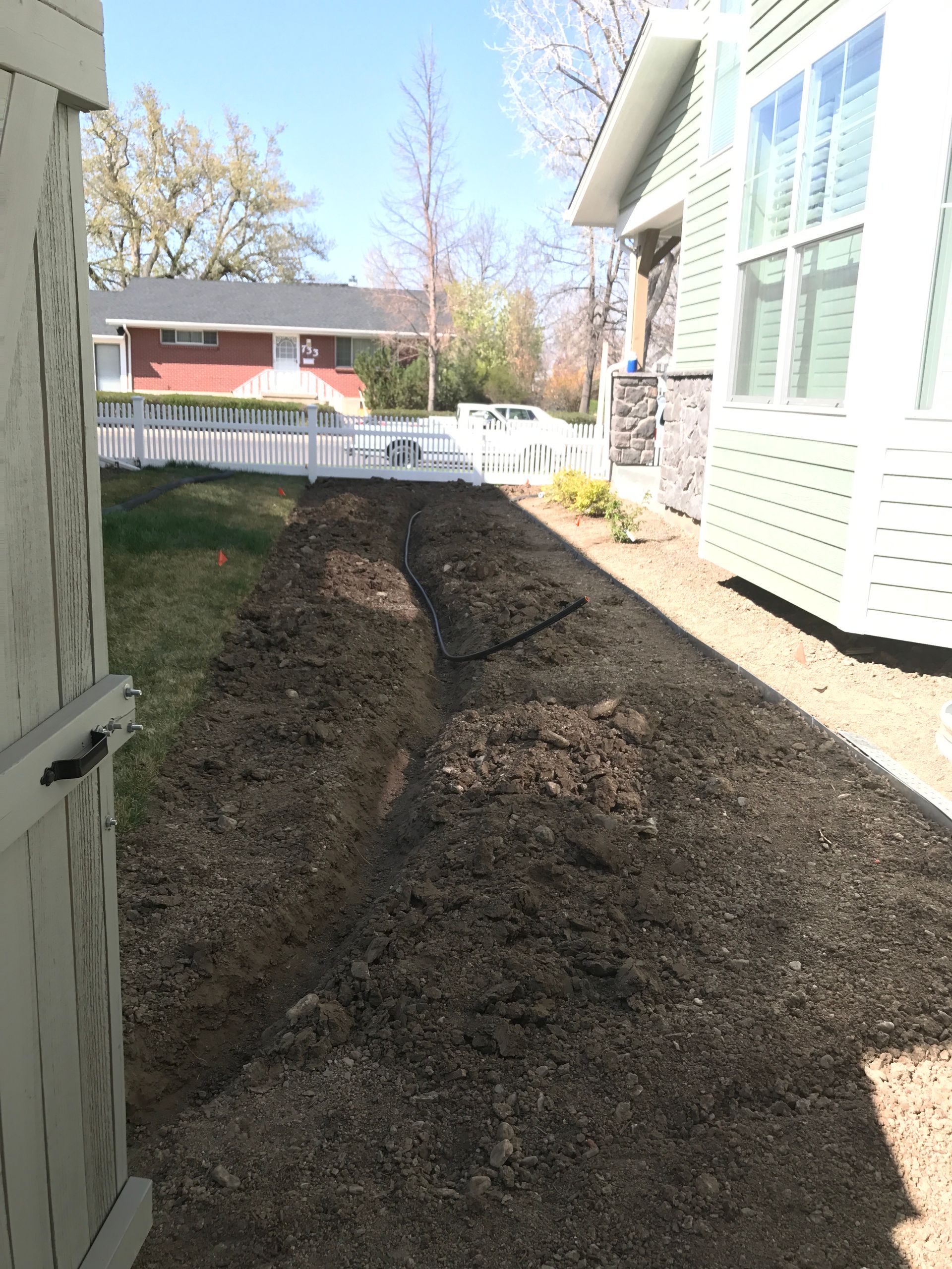 A narrow, muddy trench dug in a lawn next to a house with a white fence and a sunny sky overhead.