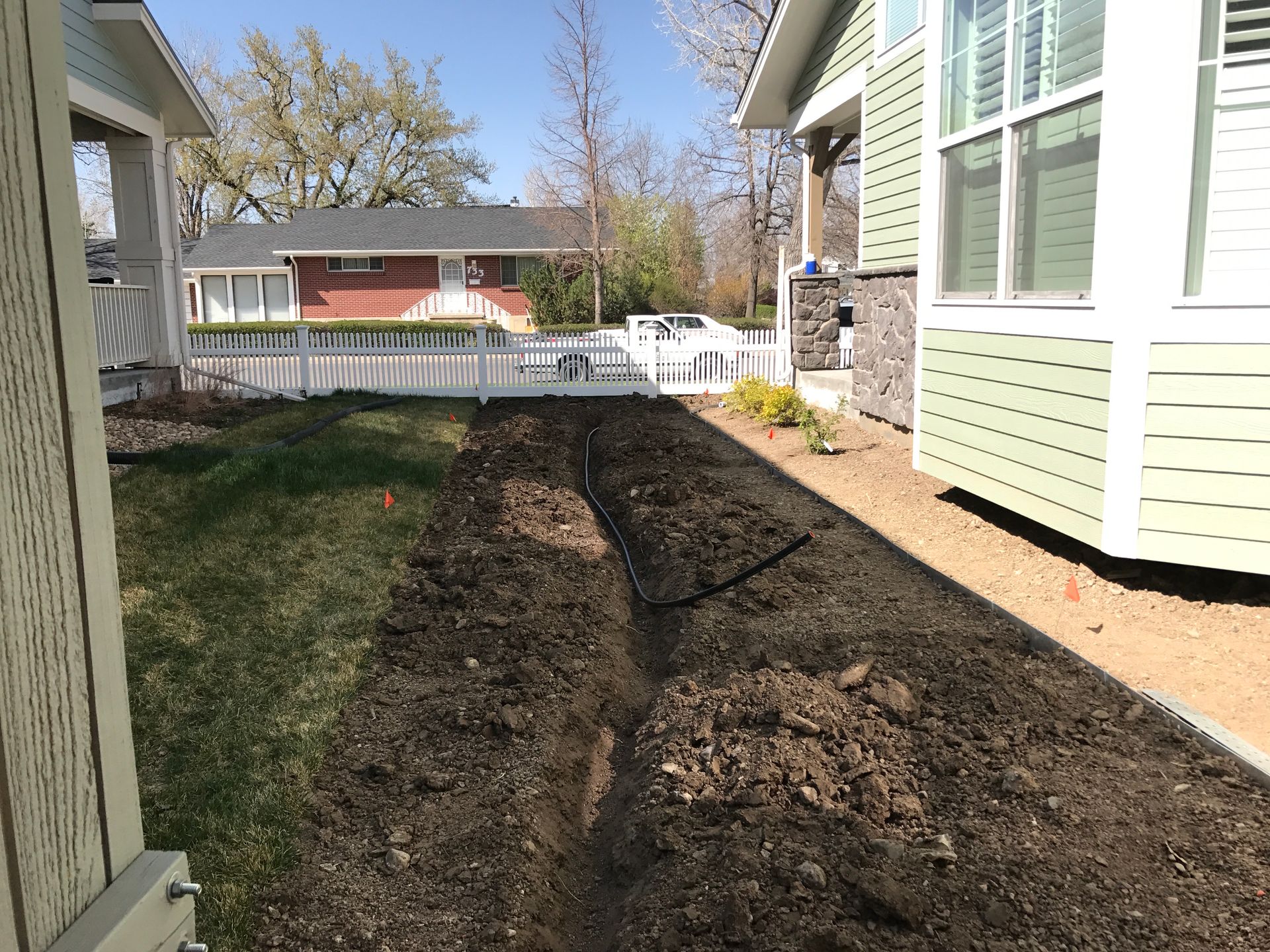 A trench dug in a yard between two houses; grass and dirt, sunny day.