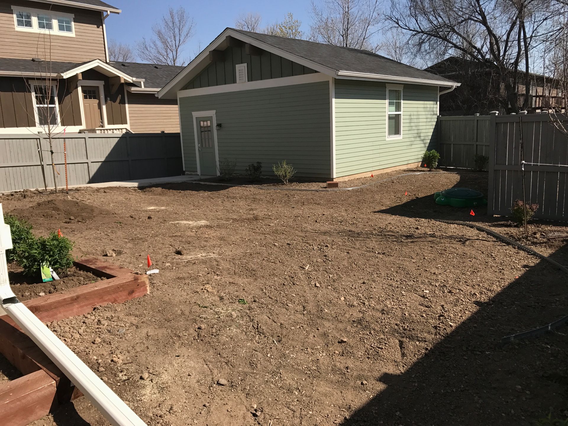 Backyard with a light green shed, brown dirt, and a wooden fence on a sunny day.