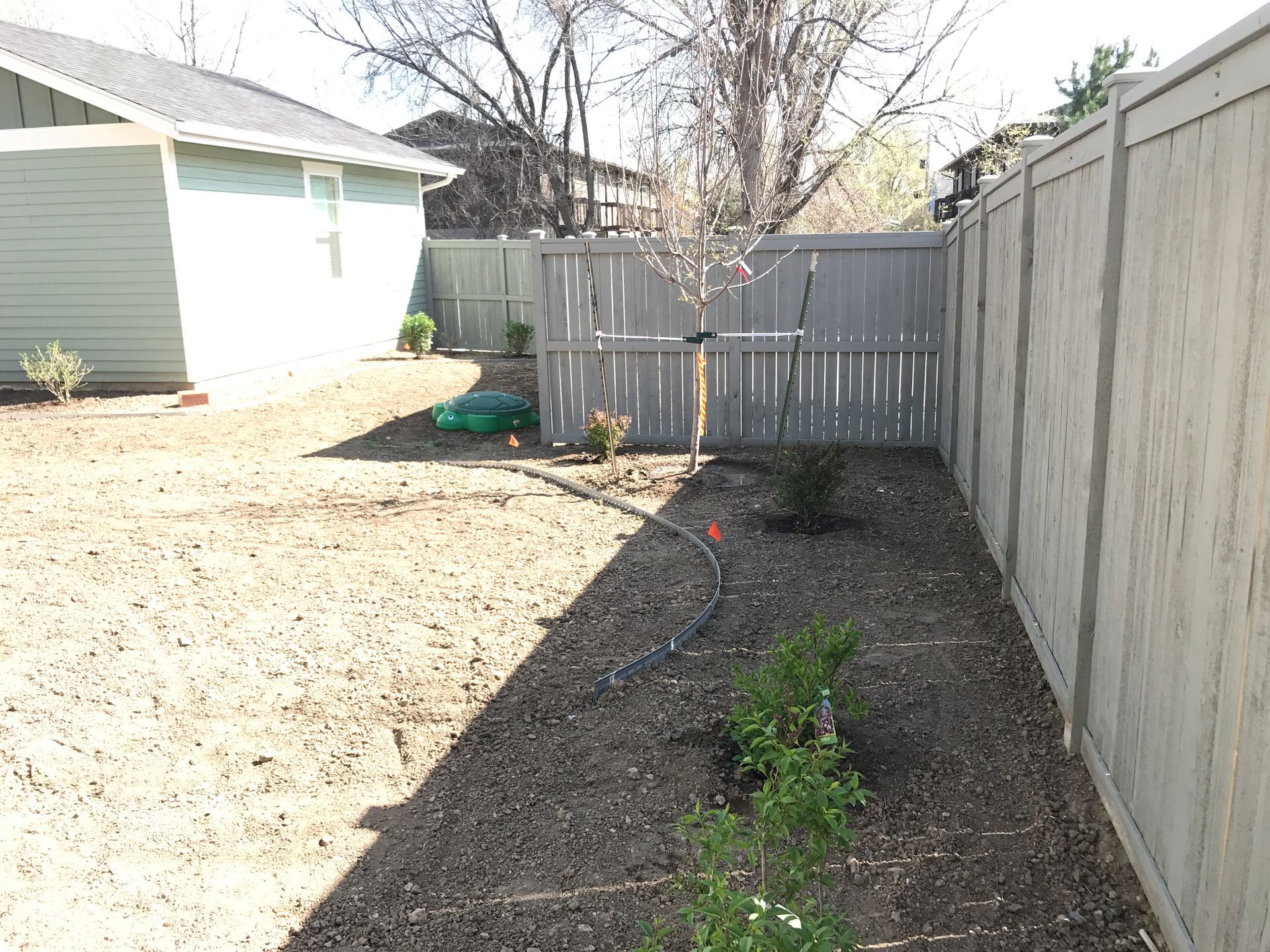 Backyard with brown mulch, green plants, a gray fence, and a light blue house.