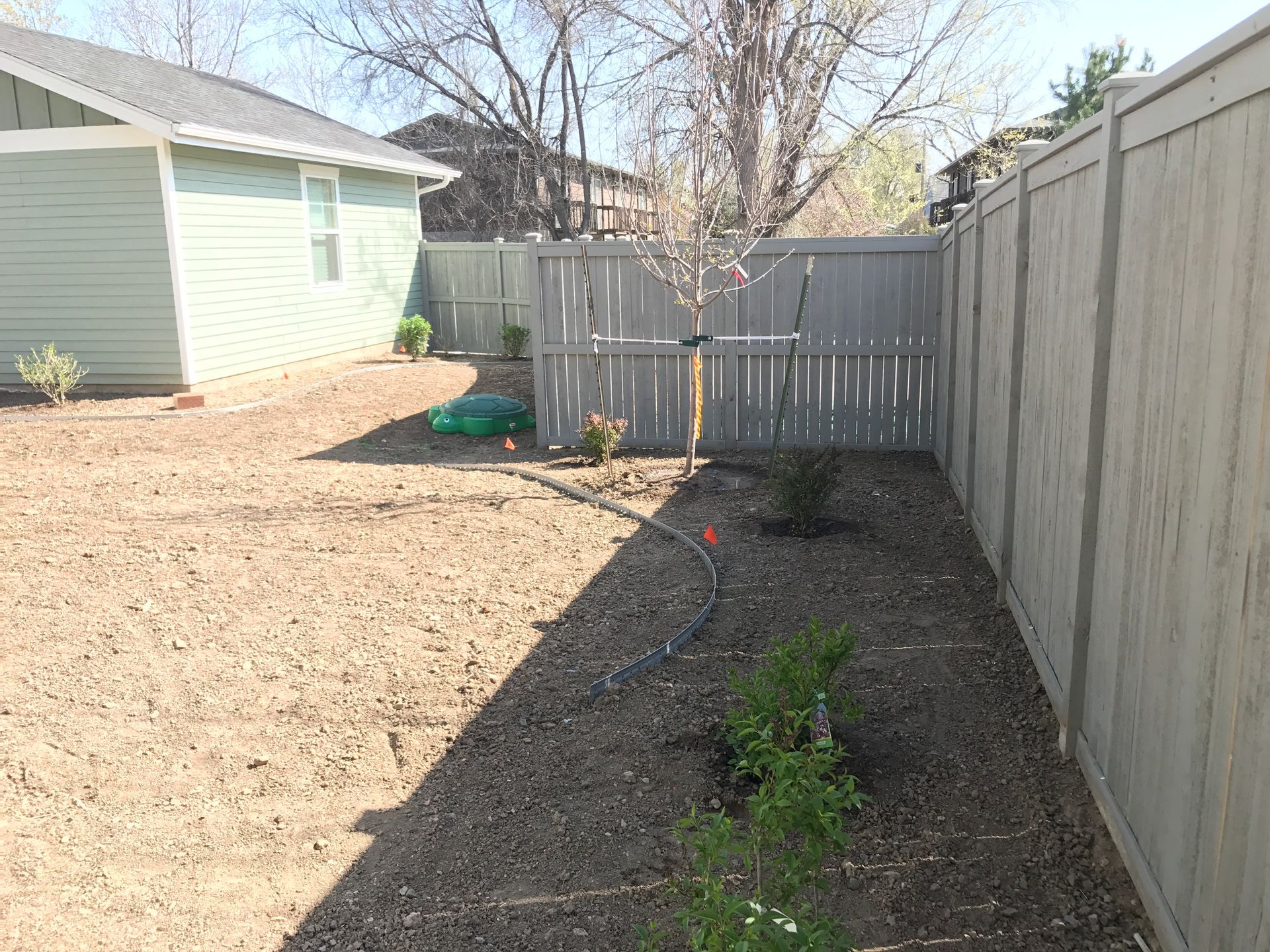Backyard with a light green house, gray fence, and dirt ground.