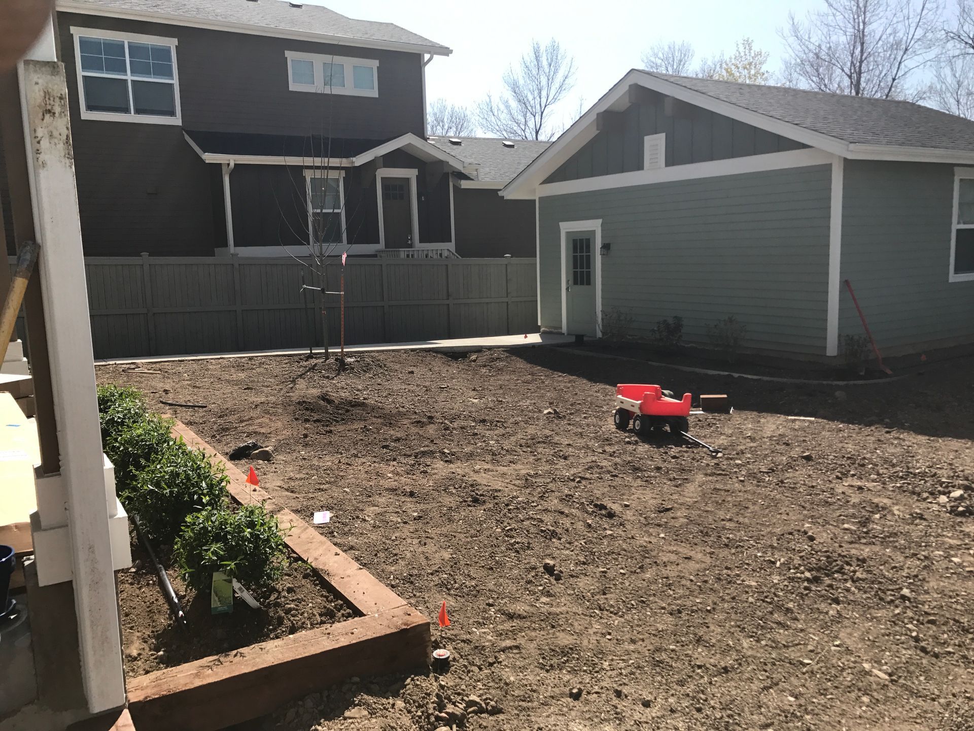 Backyard with bare soil, a small wagon, and a garage; a two-story house is in the background.