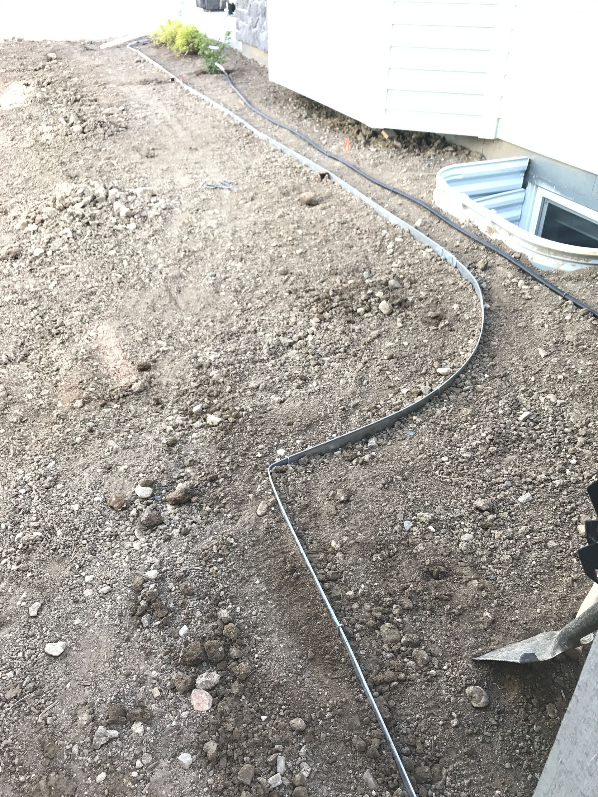 A yard with dirt and a black hose winding near a house with a window well.