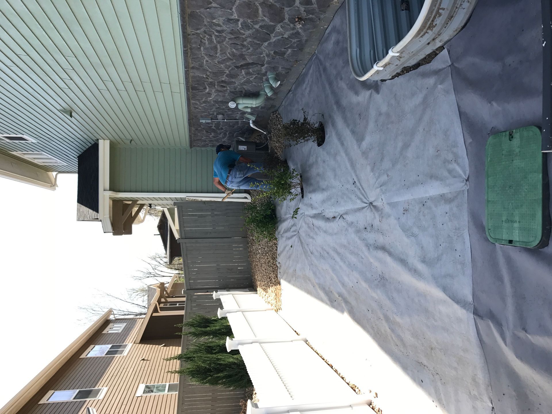Person weeding a narrow backyard with a tarp on the ground. Buildings and fence in the background.