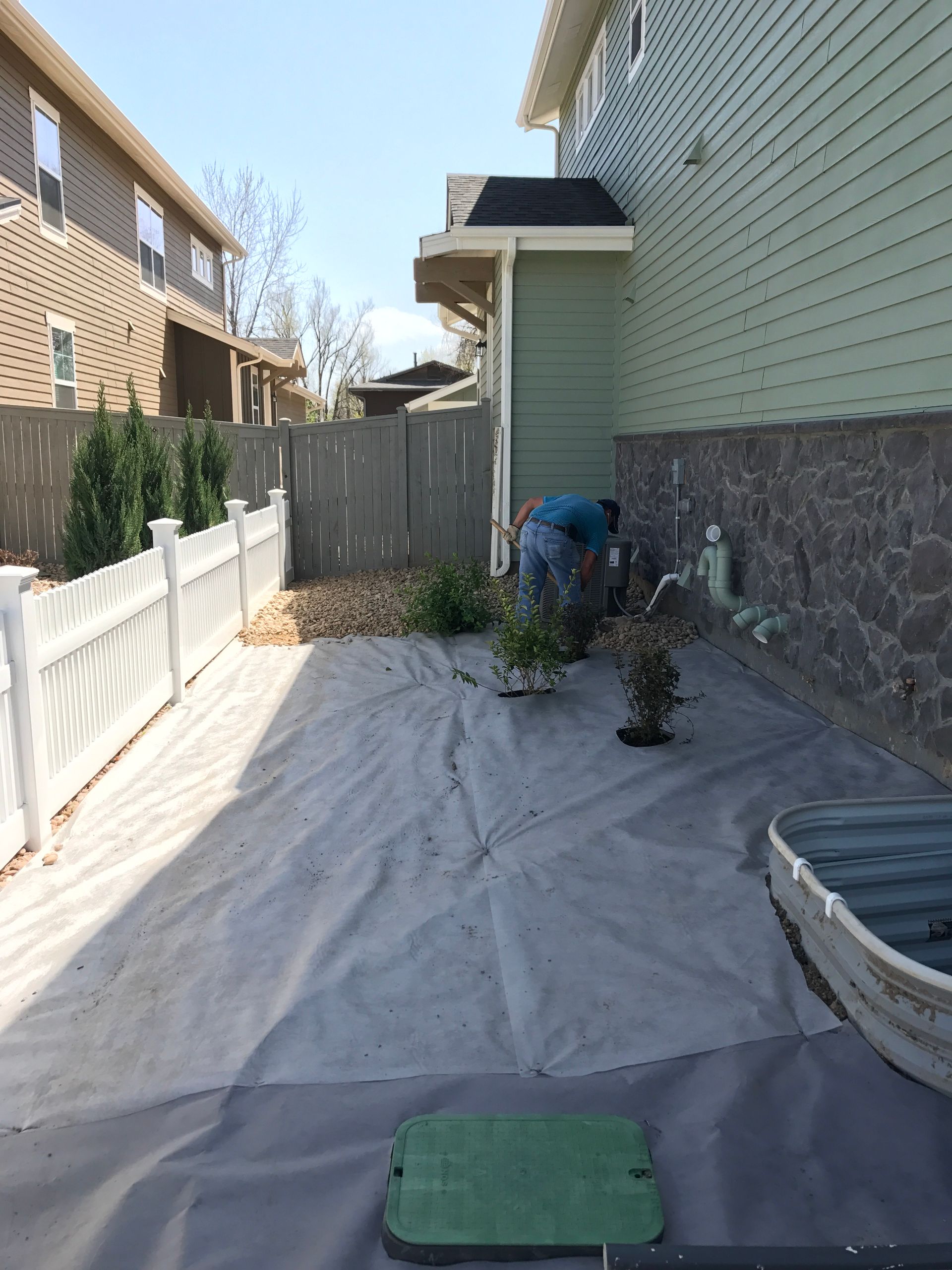 Person gardening in a backyard, covered in gray fabric. White fence on left, green house on right. Sunny day.