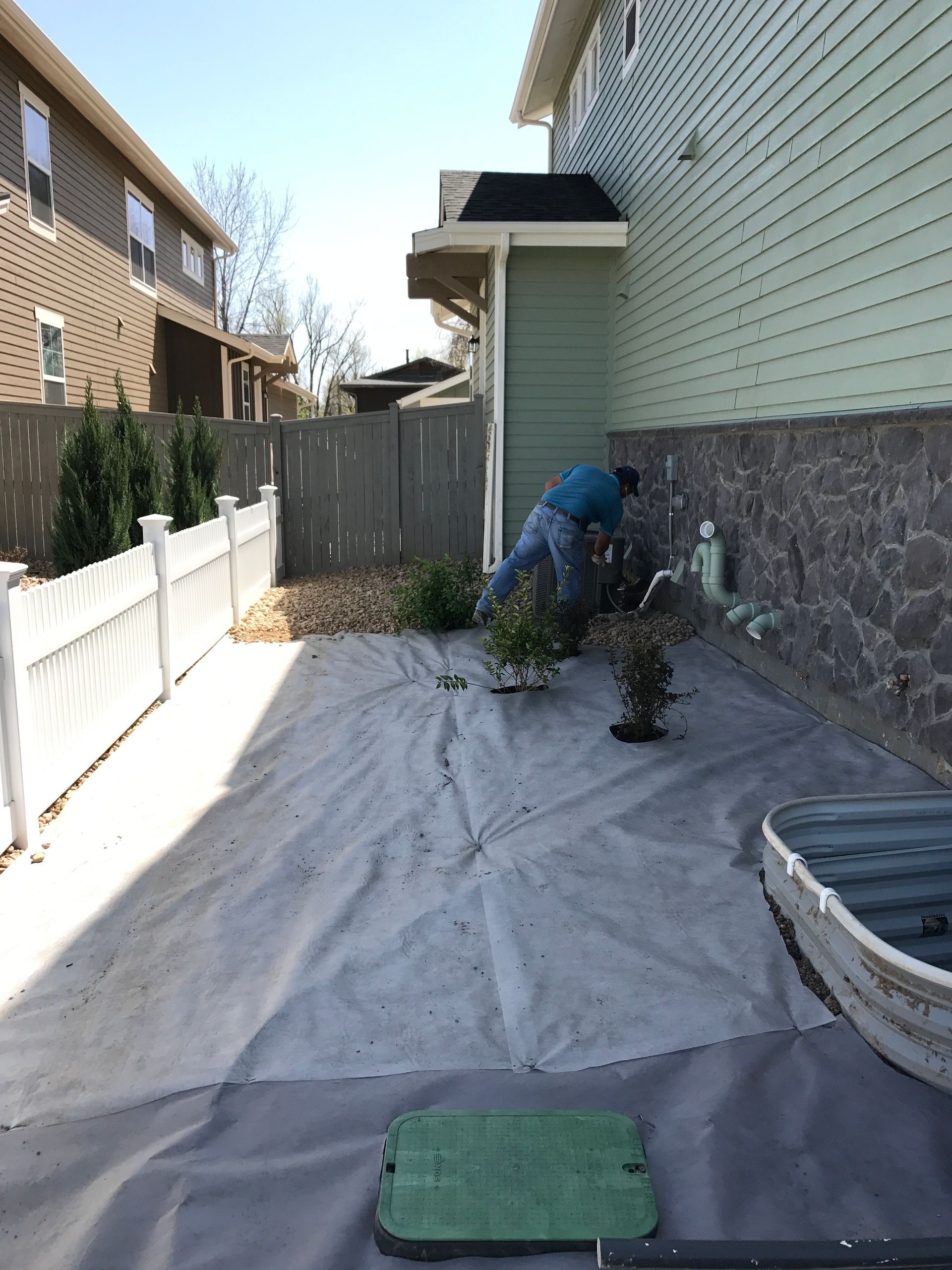 Man tending small plants in a backyard with a gray ground cover. A boat is to the right.