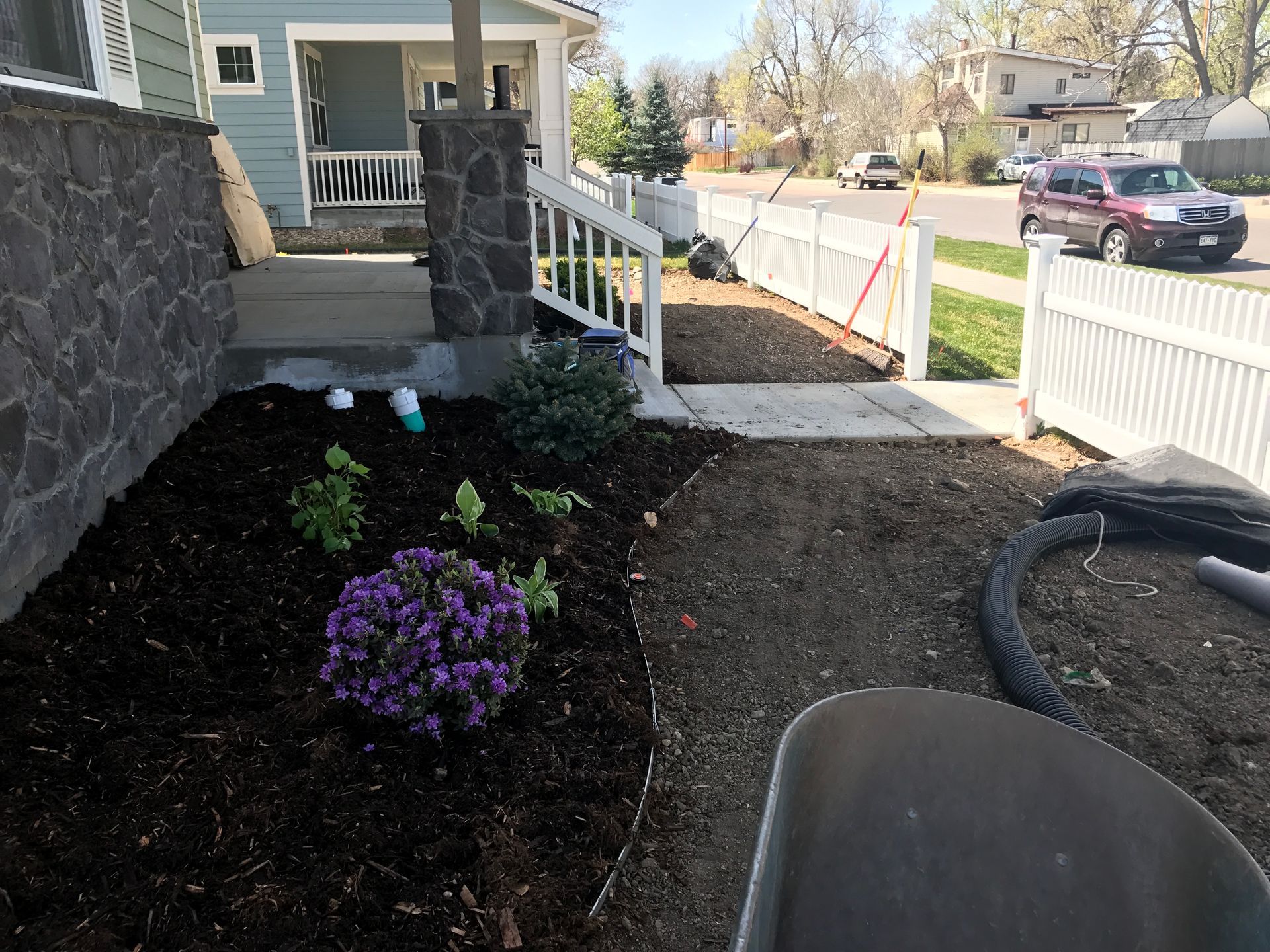 A front yard with dark mulch, plants, white fence, and a teal house. A car drives by.
