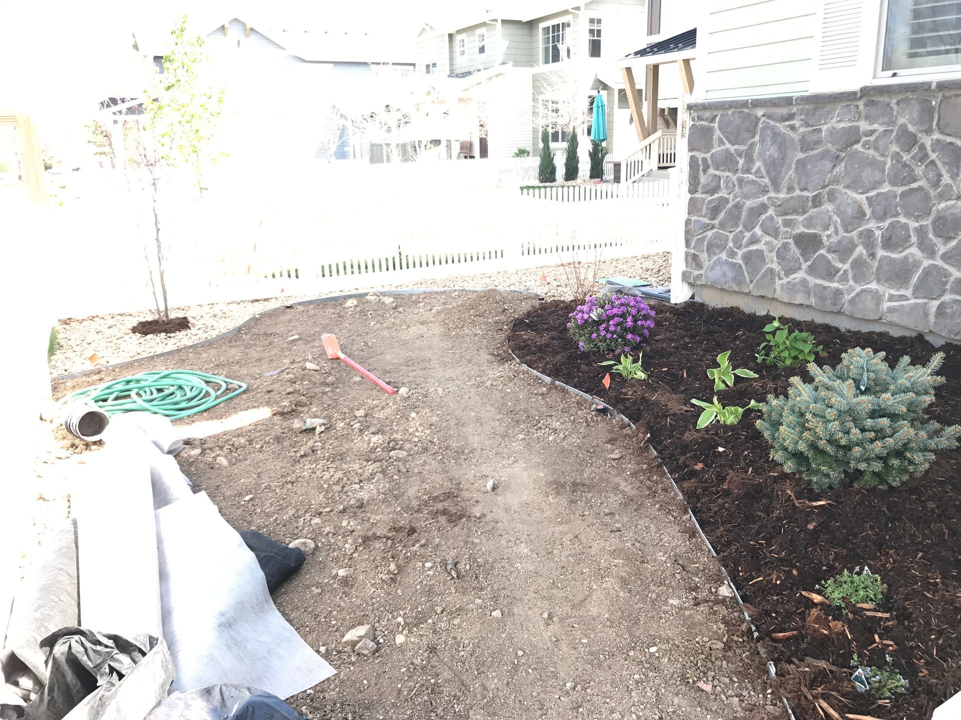A gravel path curves through a front yard, with a stone wall and recently planted flowers.