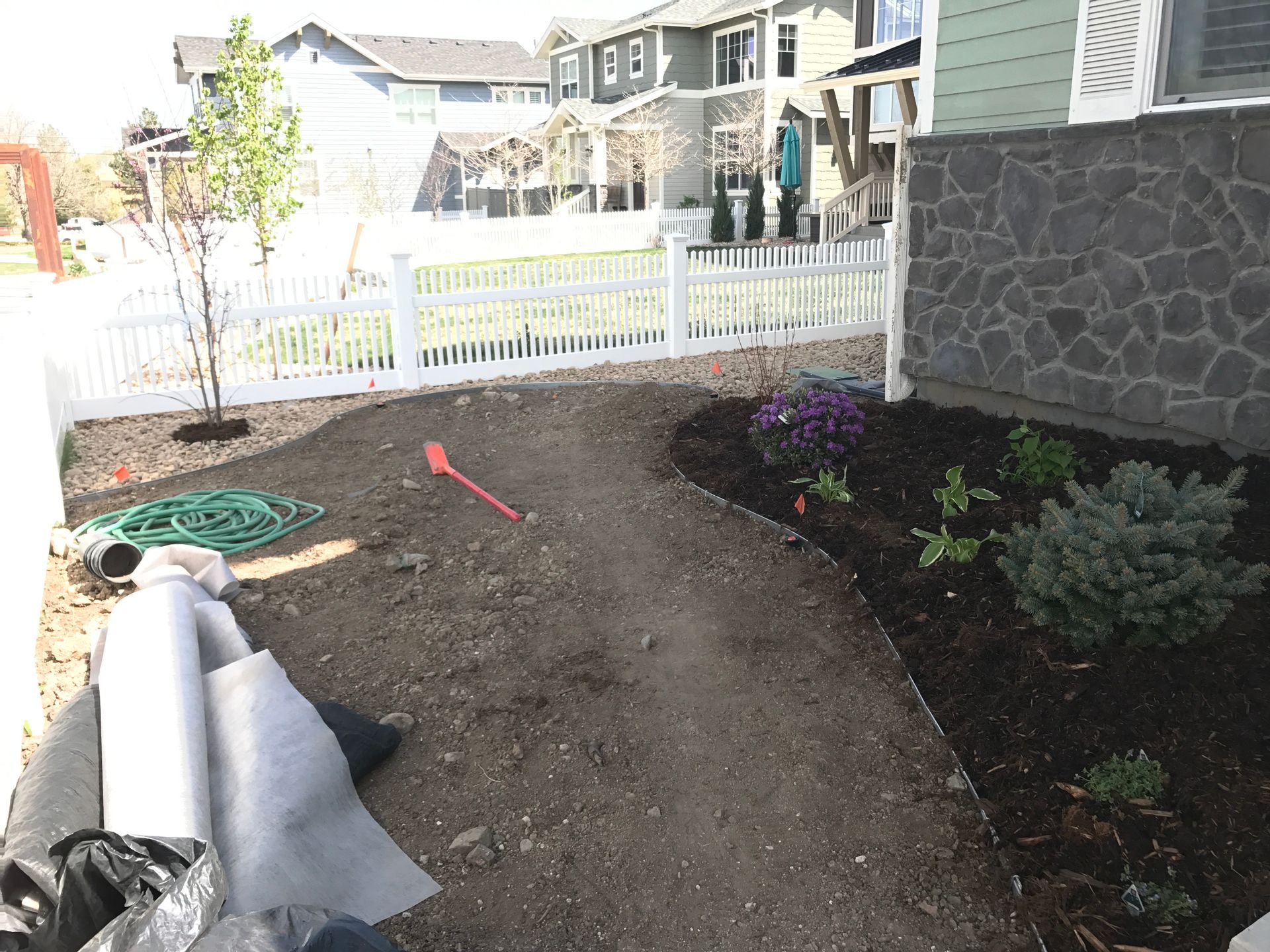 A dirt pathway curves through a front yard, edged with dark mulch and plants, leading to a white picket fence.