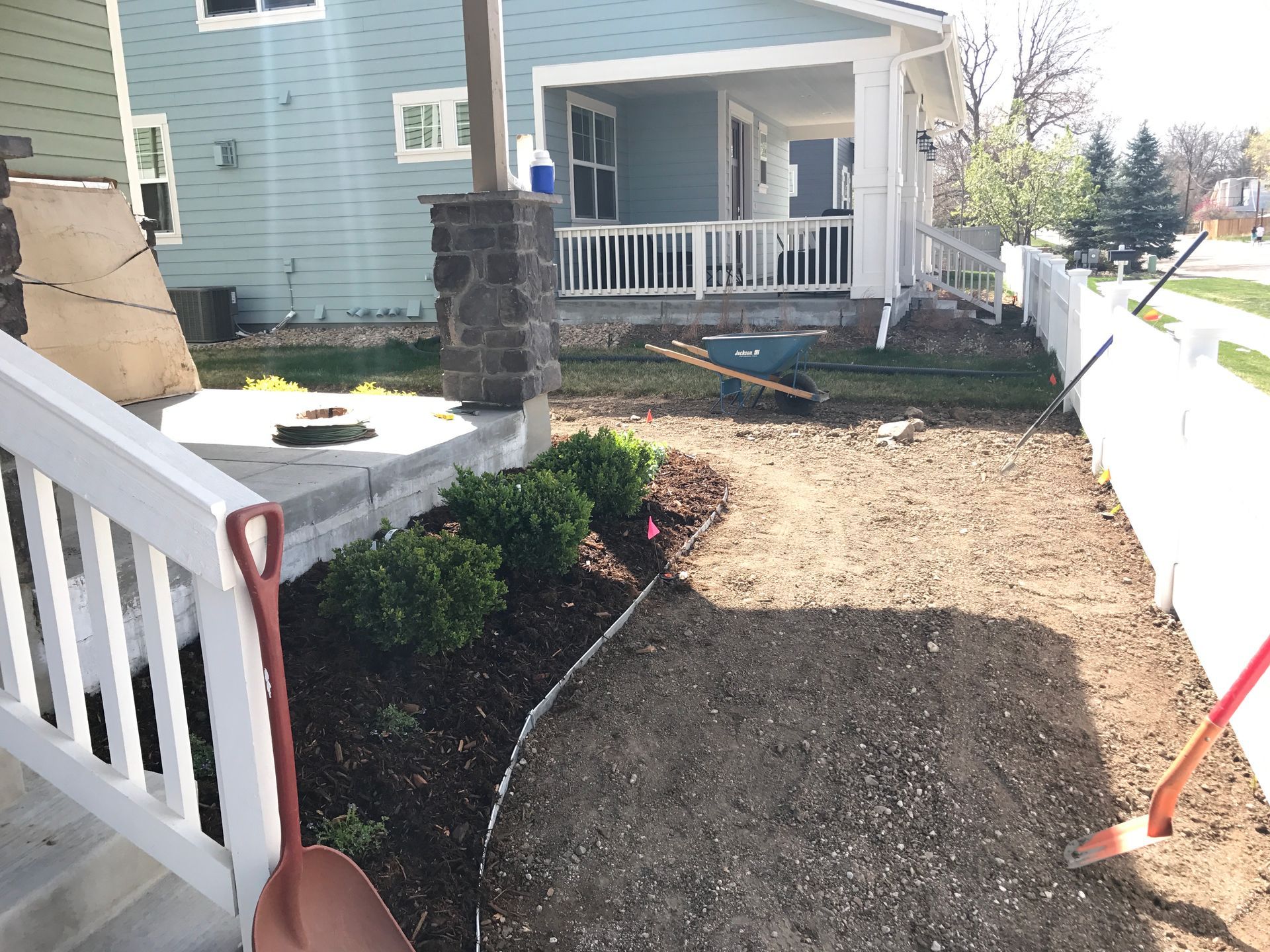 A front yard with landscaping under construction: dirt path, bushes, fence, house, and tools.